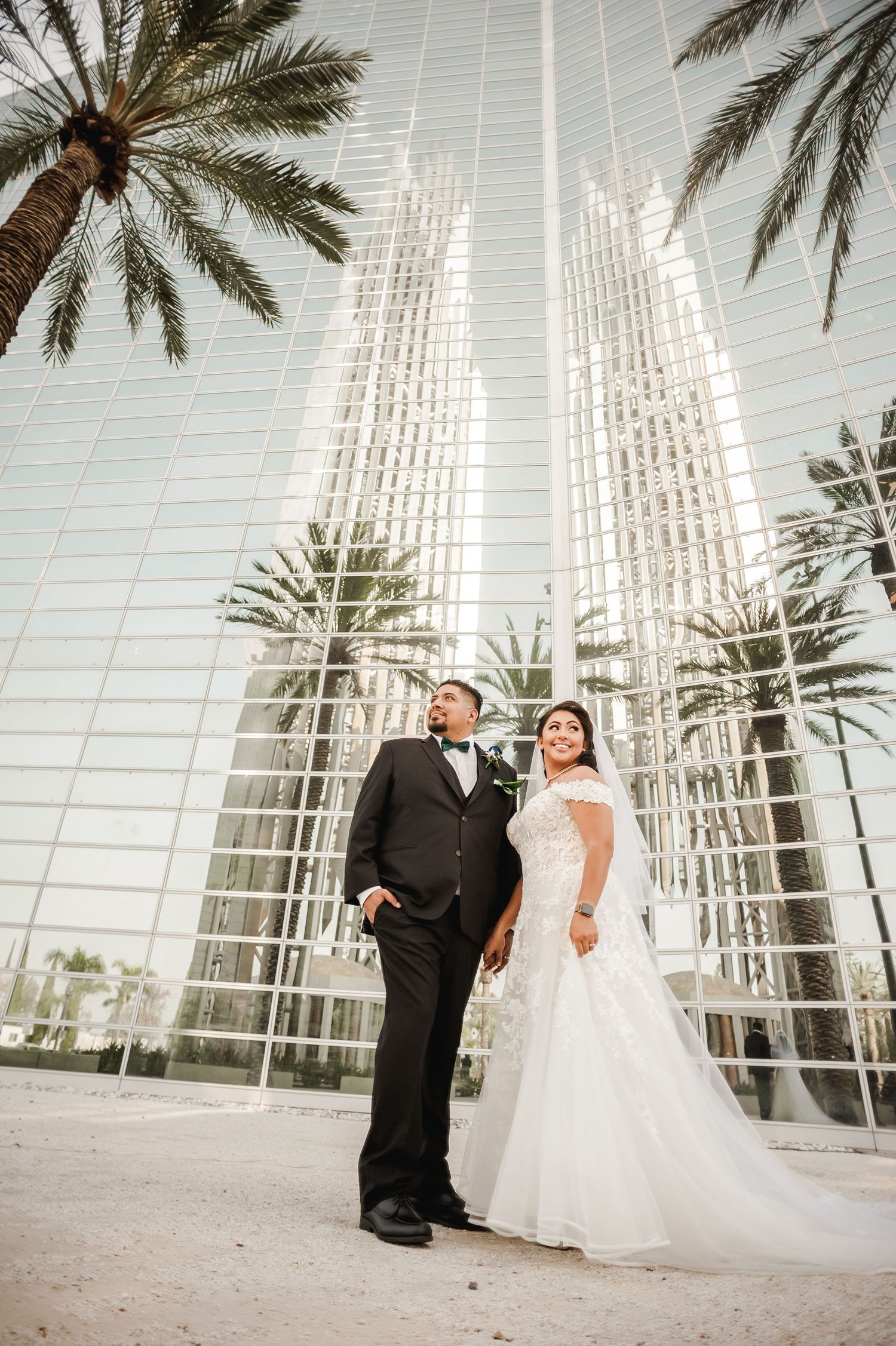 Elegant wedding photo with couple standing by palm trees and glass architecture.