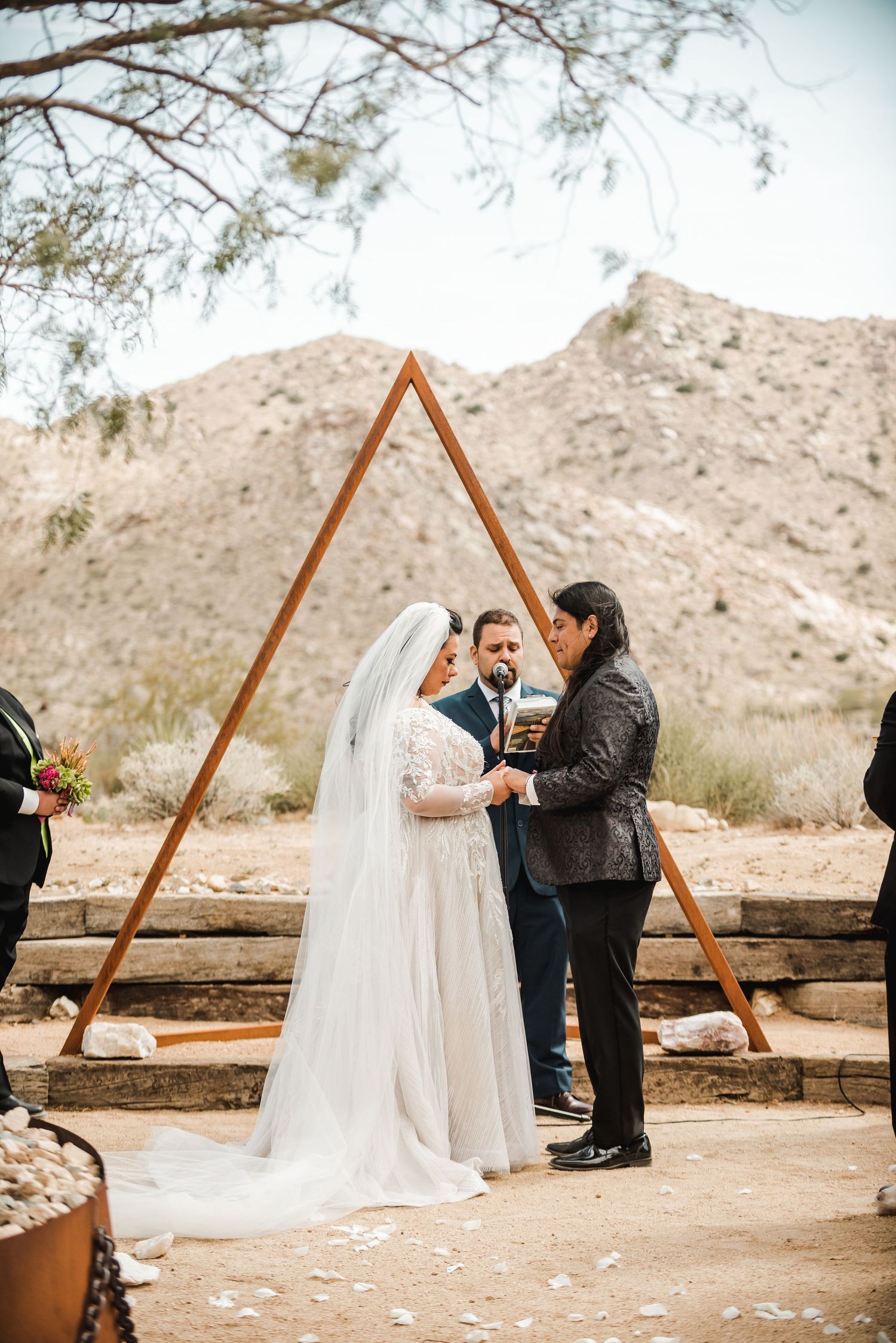 A wedding ceremony takes place under a wooden triangle arch in the desert with mountains in the background.