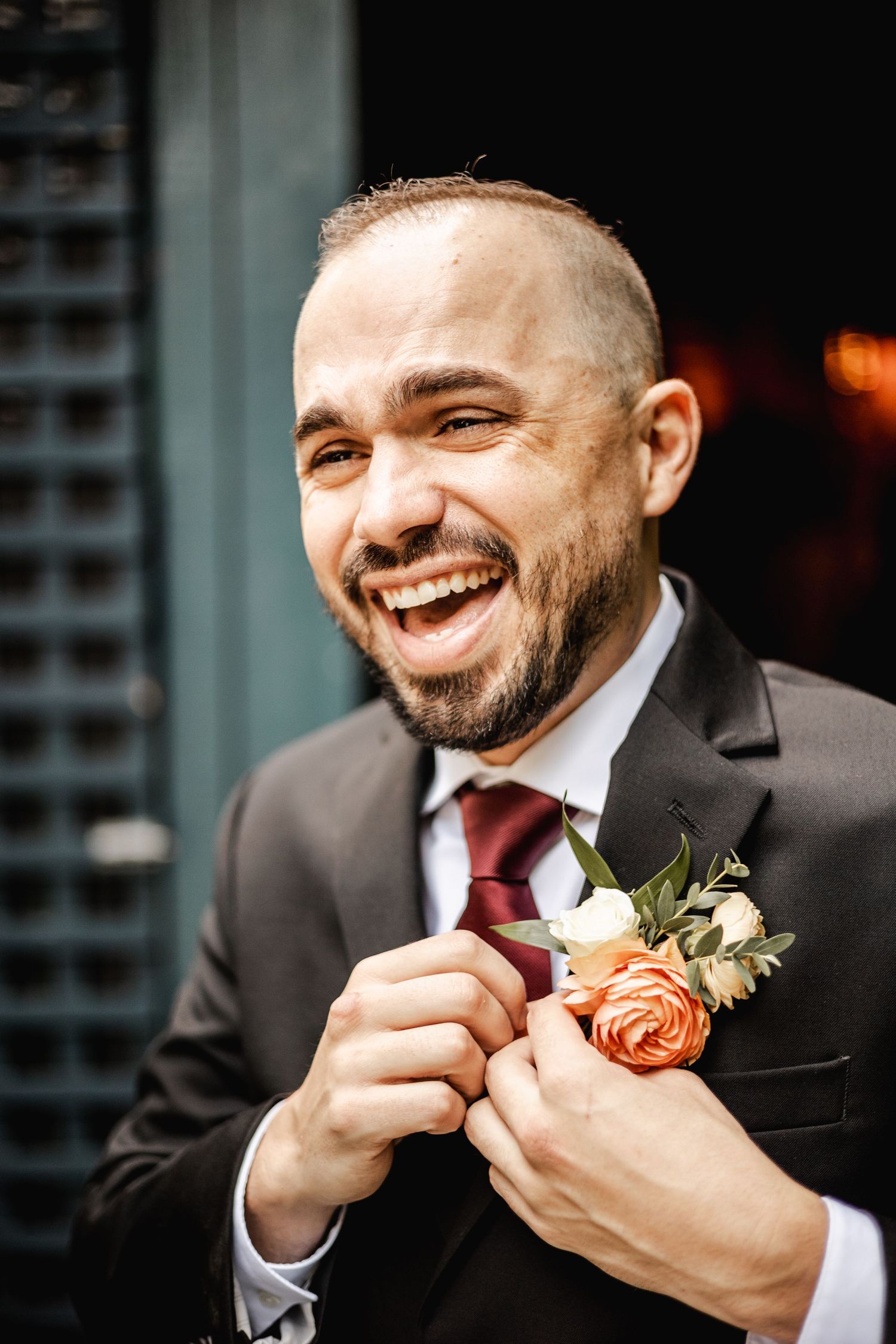 A groom in a black suit and burgundy tie laughs while adjusting his coral boutonniere during a wedding preparation moment.