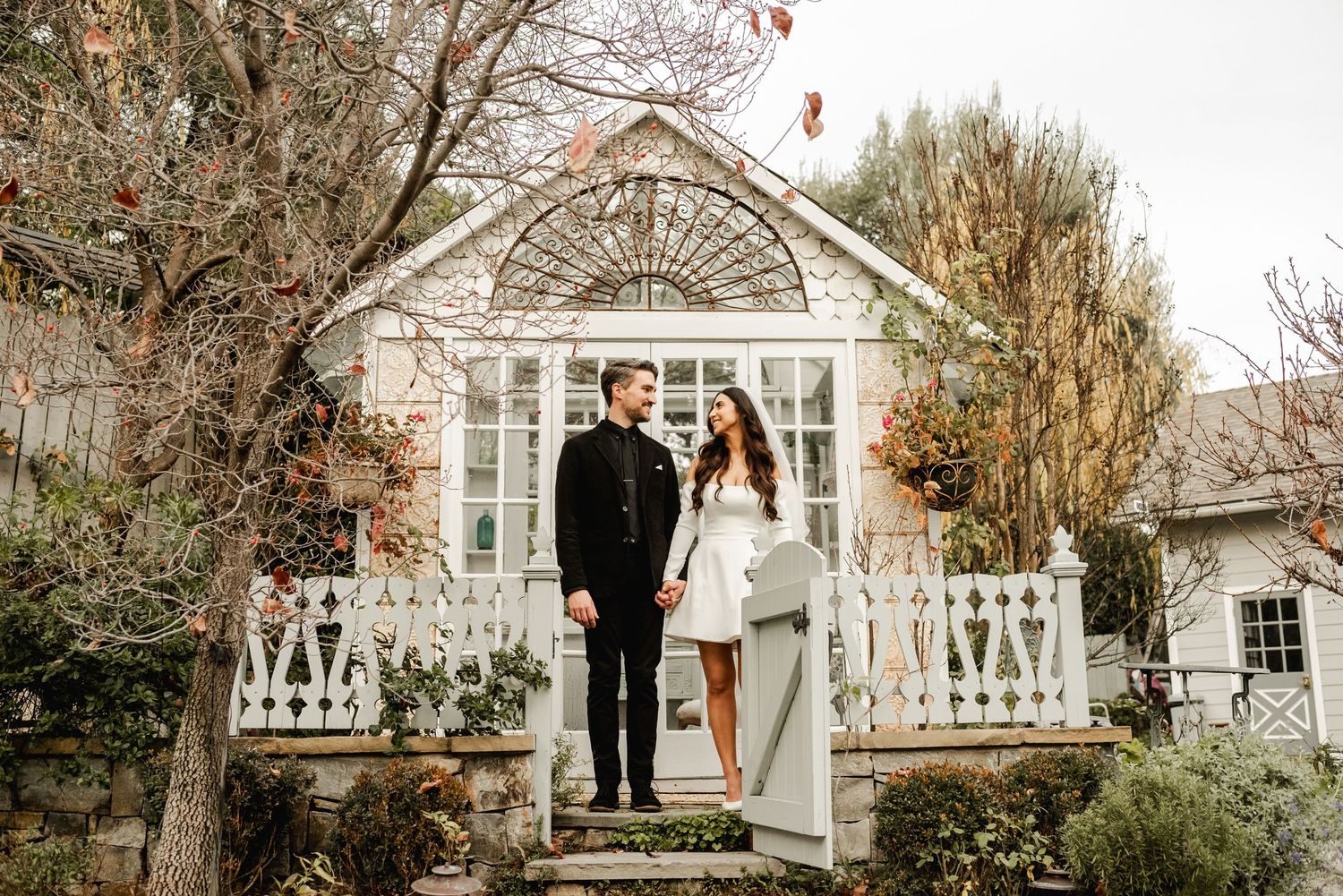 A couple stands on the steps of a charming white Victorian cottage with ornate trim on their wedding day.