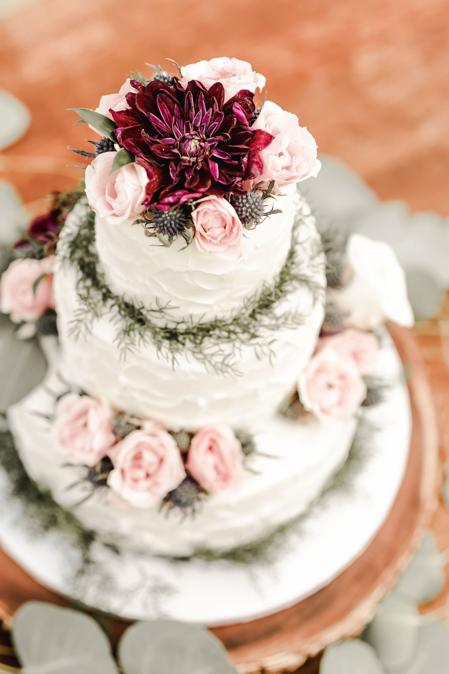 White wedding cake decorated with burgundy dahlias and pink roses on copper charger.