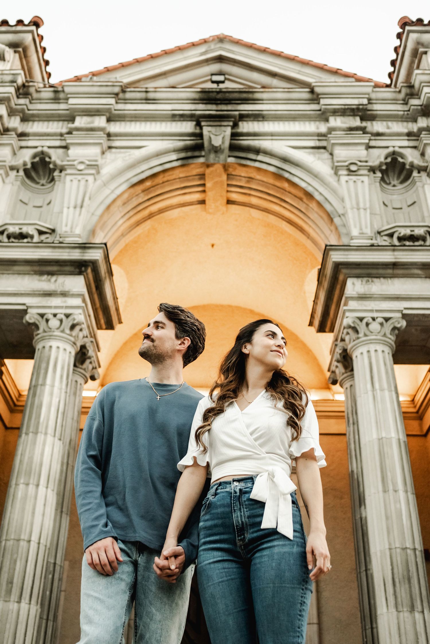 Couple in casual attire holding hands under ornate stone archways and columns.