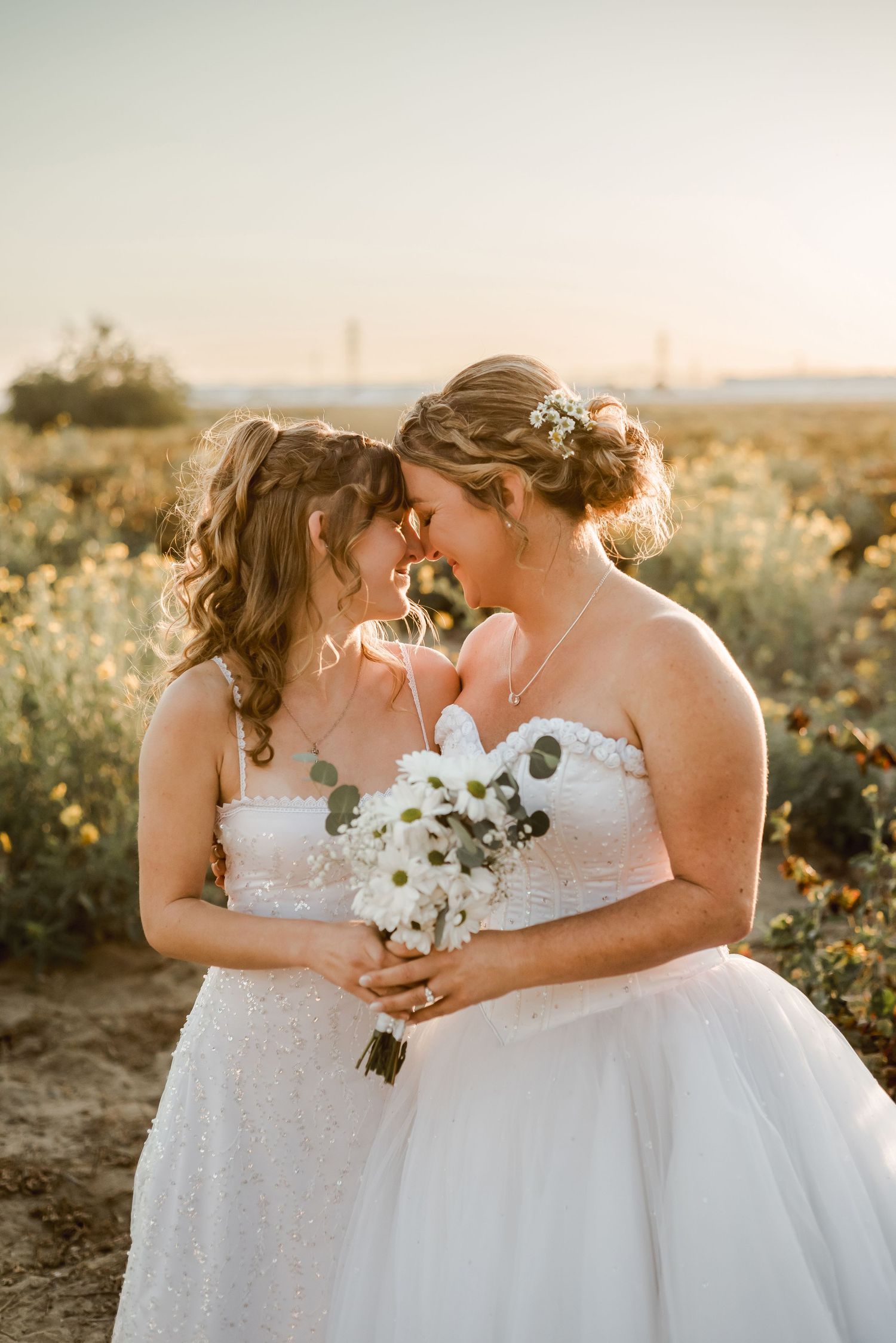Two brides sharing an intimate moment while holding white floral bouquets at sunset.