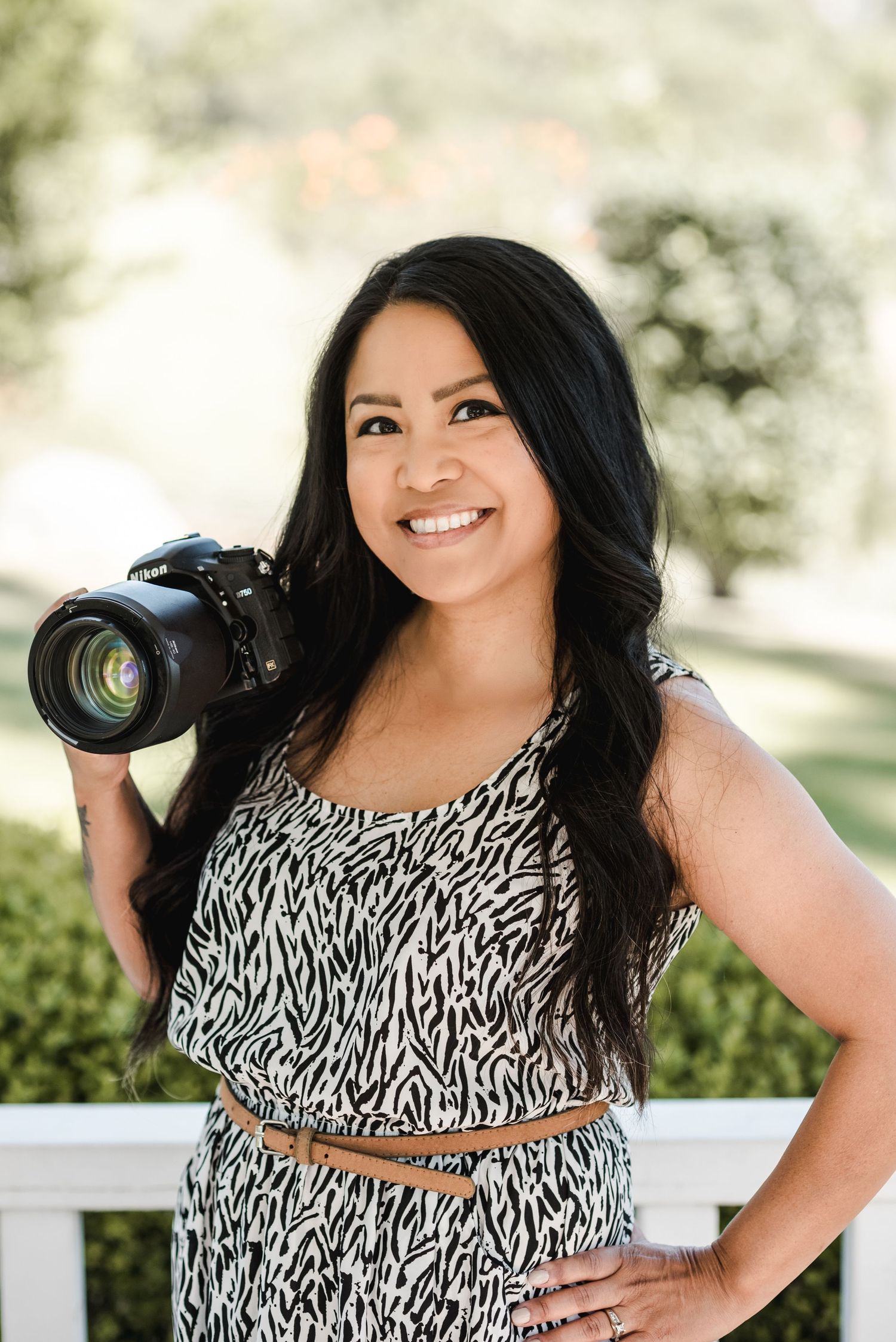 Professional female photographer in black and white patterned dress holding camera while smiling outdoors.