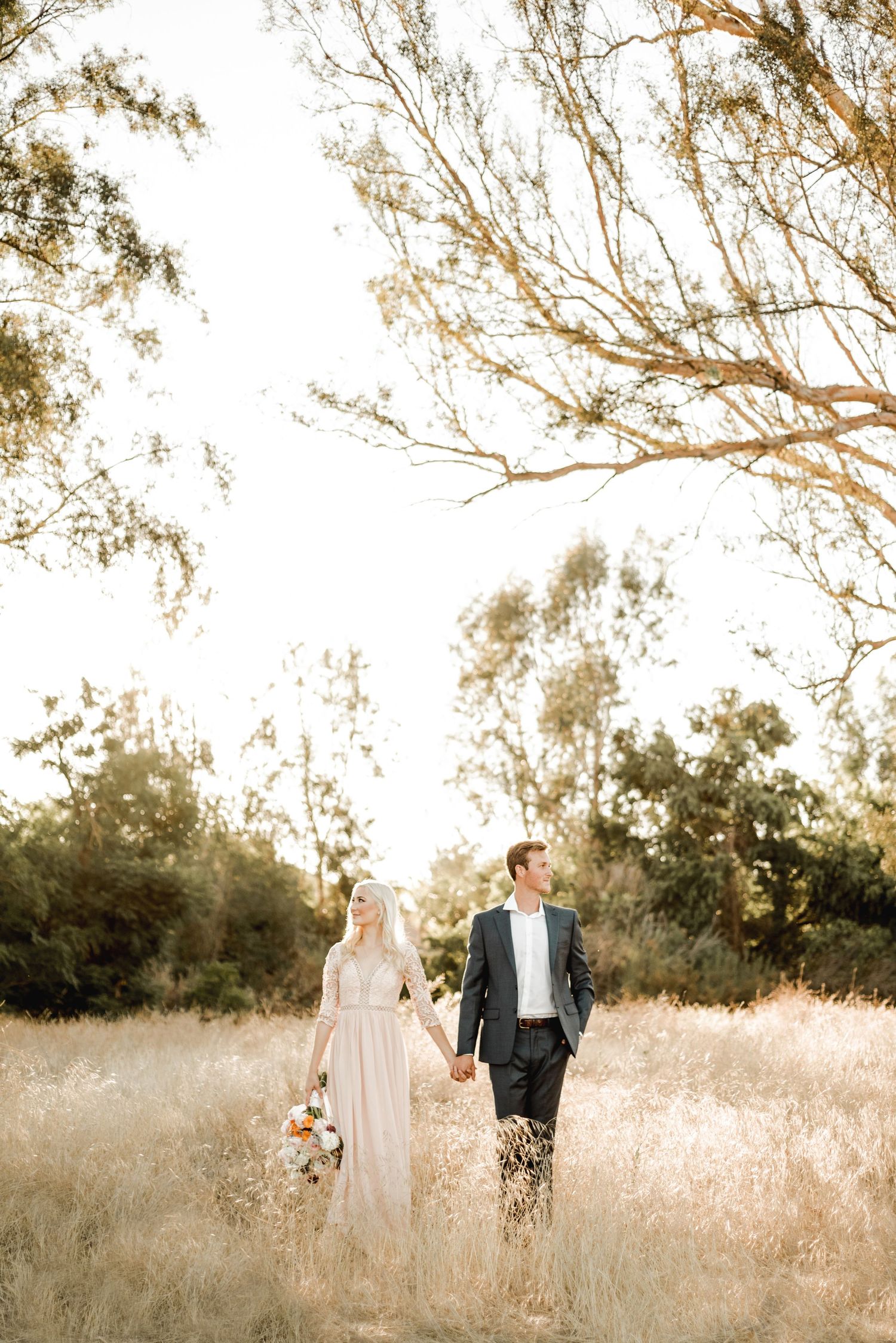 Couple walking through sun-drenched field with eucalyptus trees in background.