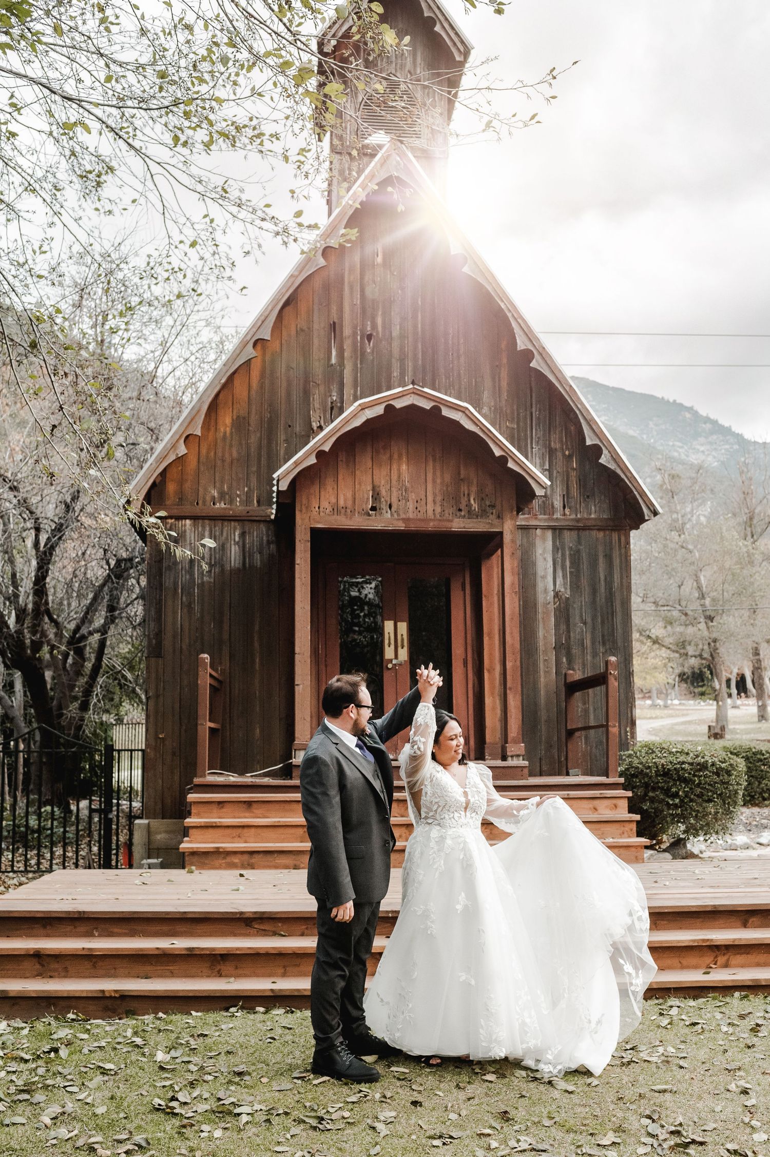 A couple celebrates outside a rustic wooden chapel with sunlight streaming through the trees.