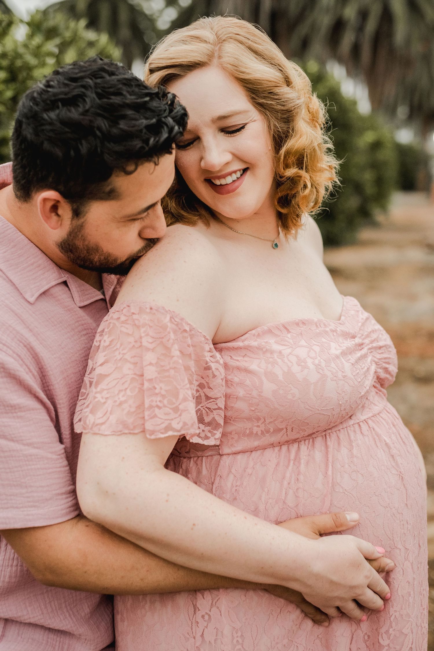 An intimate maternity session featuring a couple in pink outfits embracing in front of palm trees.