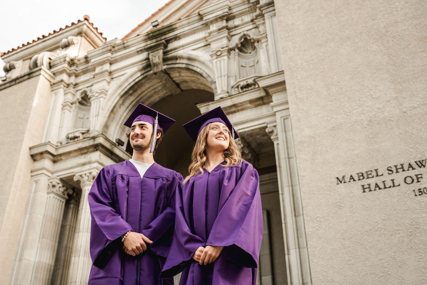 Graduates in purple robes pose in front of a historic stone building with ornate architecture.