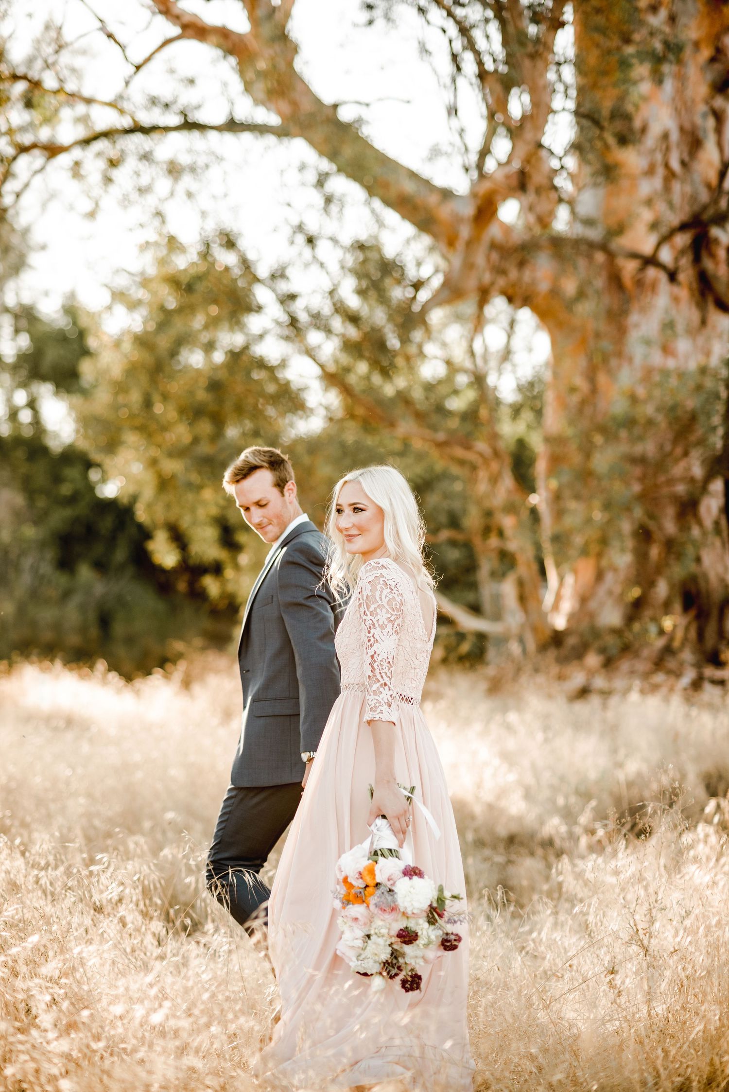 Couple in formal wedding attire pose back-to-back in golden field with bouquet.