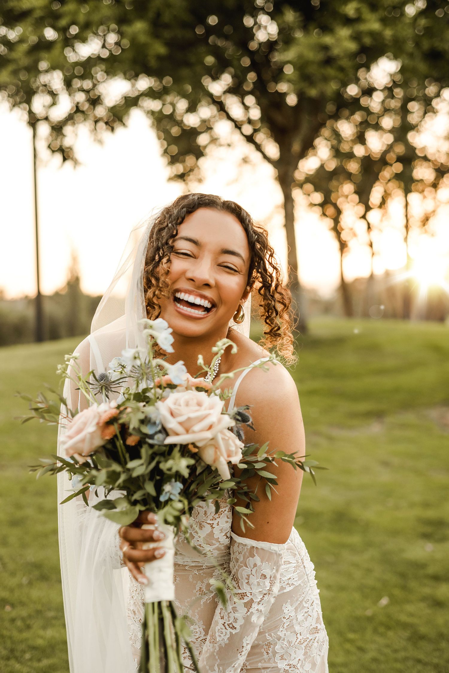 Joyful bride laughing while holding white floral bouquet in outdoor garden setting.
