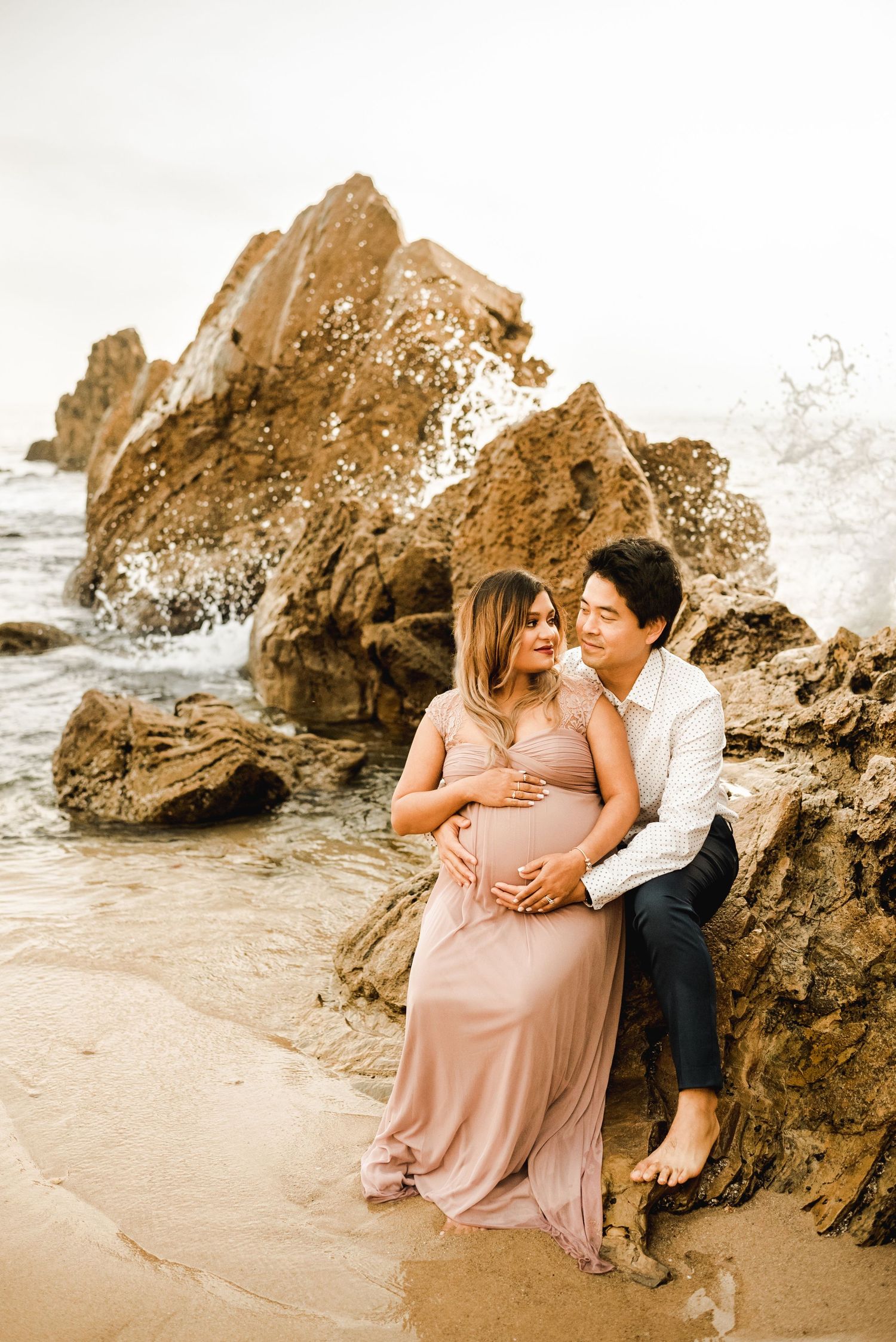 A couple shares intimate moments on a beach with dramatic rock formations at sunset.