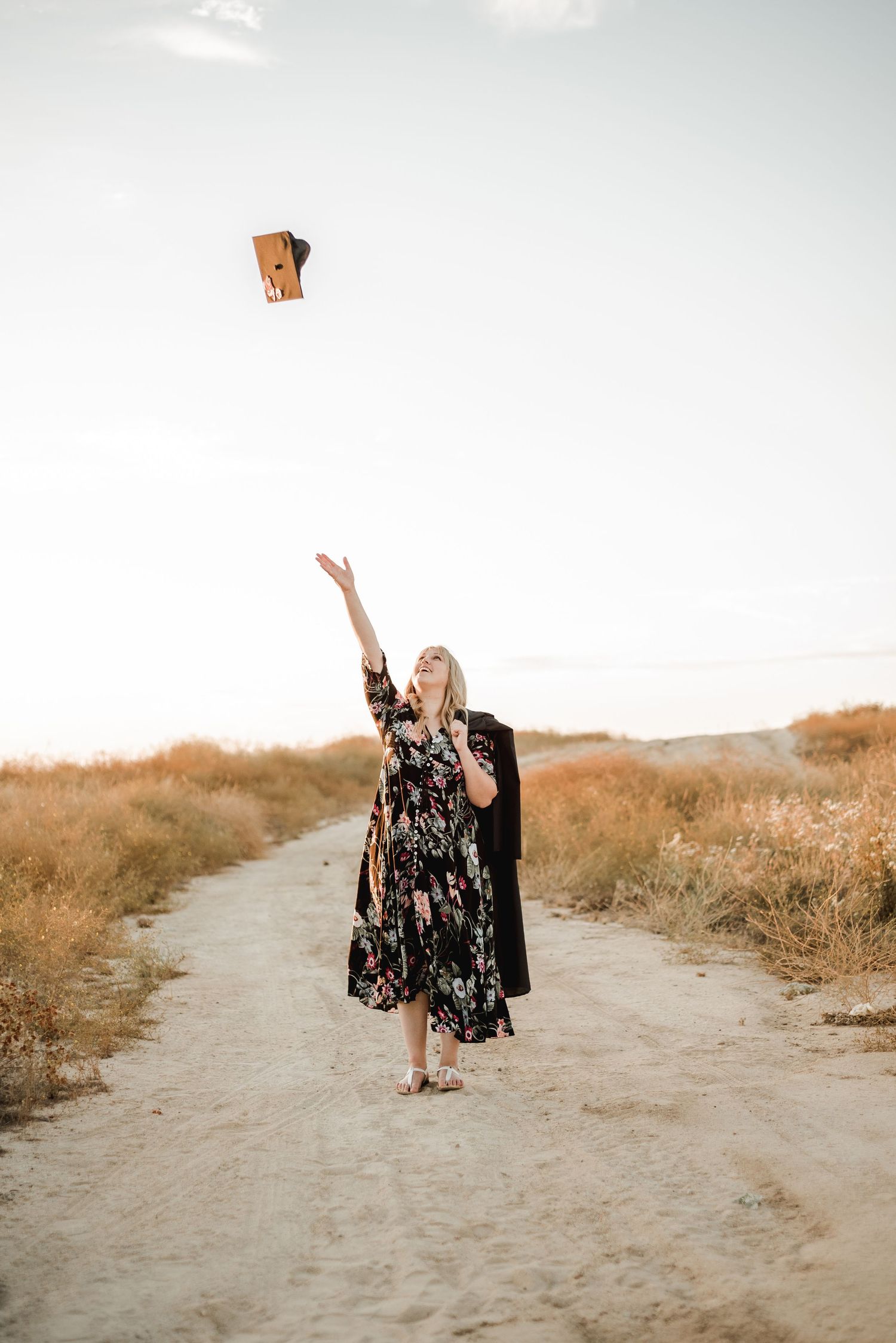 A floral dress-clad figure tosses a graduation cap against a golden sunset on a dirt path.