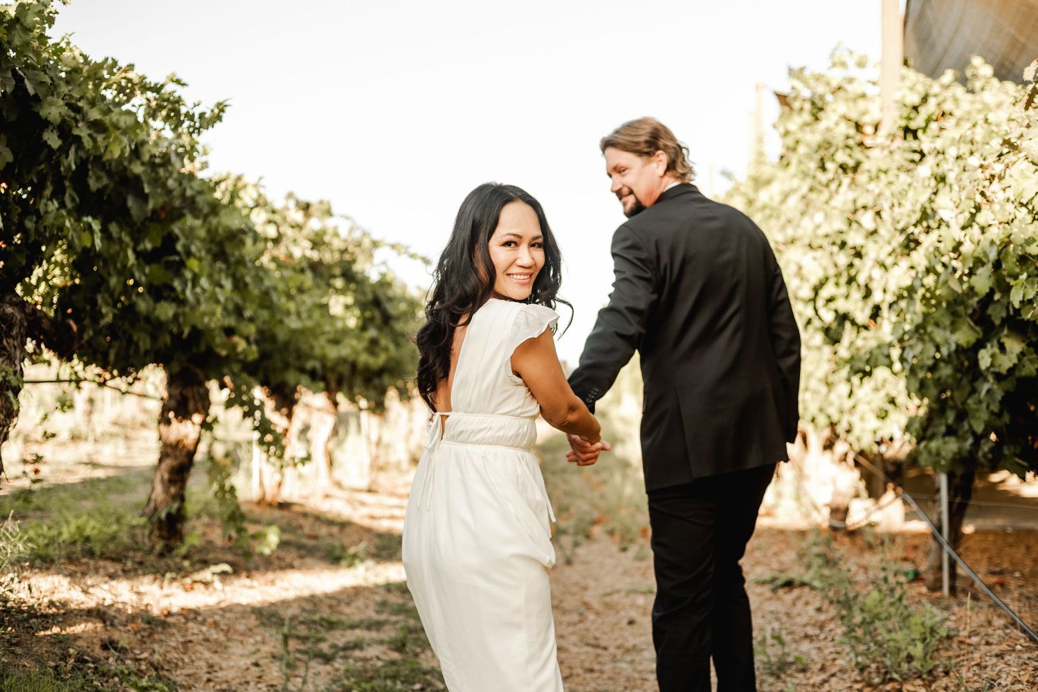A couple walks through a sunny vineyard during their wedding photoshoot.