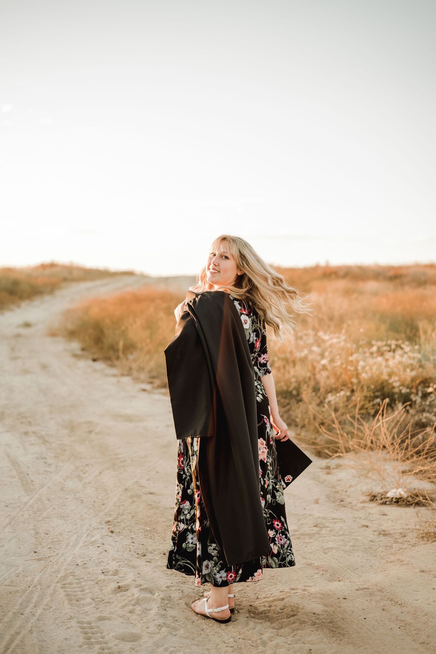 A floral dress and graduation gown flow in the wind on a rural path at sunset.
