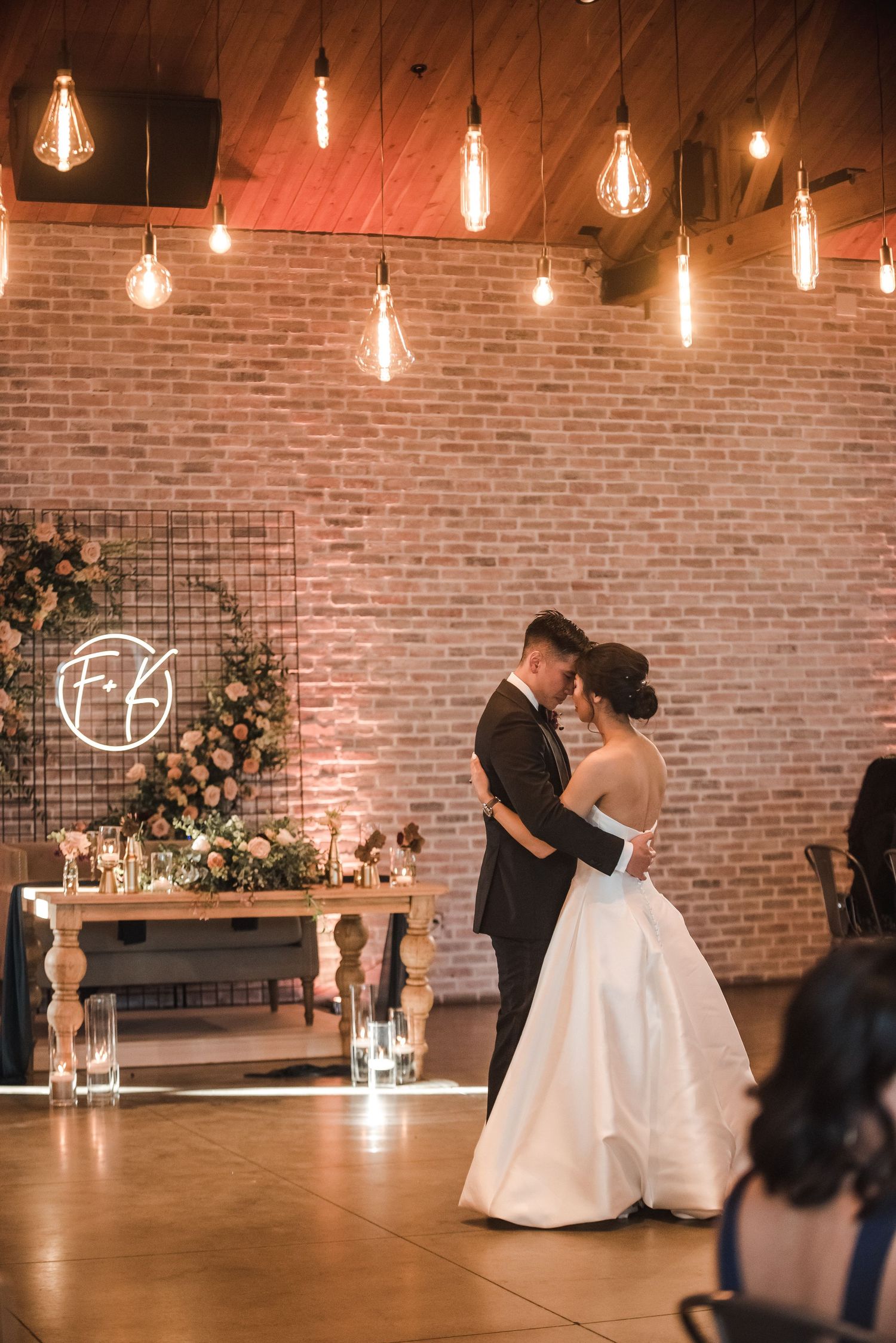 A romantic wedding reception scene with vintage Edison bulbs hanging above a couple dancing near a decorated brick wall.