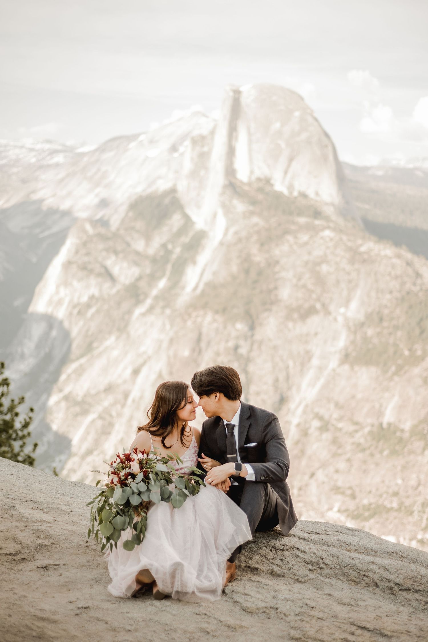 A couple sits together on a mountain ledge overlooking Half Dome in Yosemite National Park.