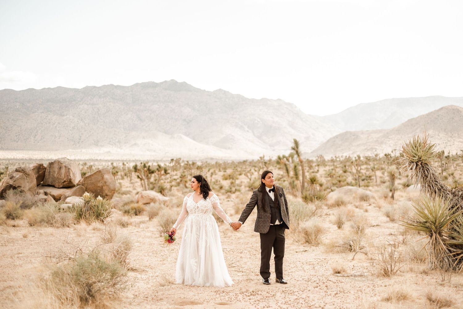 A couple walks through a desert landscape with mountains in the background.