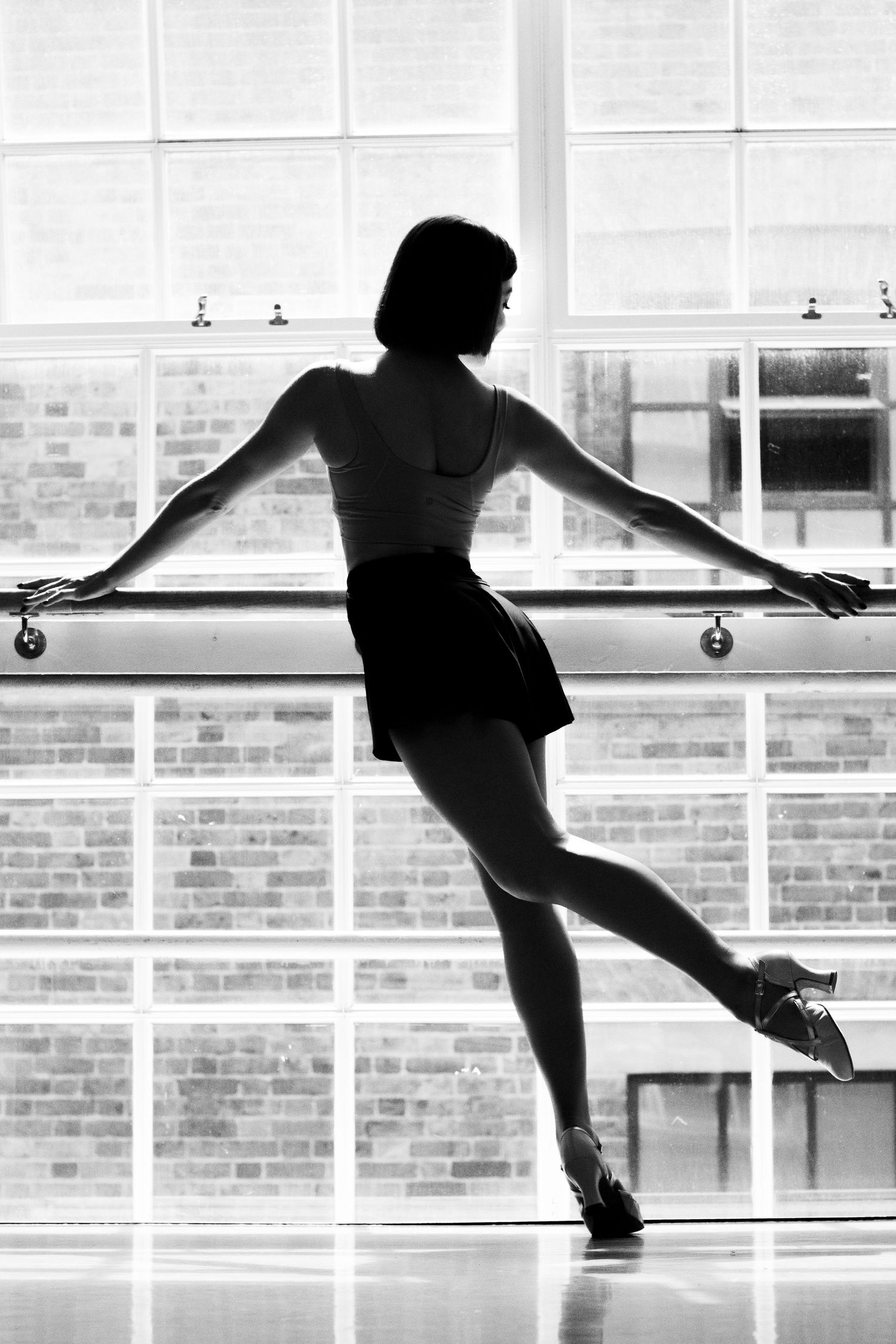 Black and white image of one of a performer modelling a shoe from LaDuca Shoes. In front of a large window in a dance pose, showing the shoe