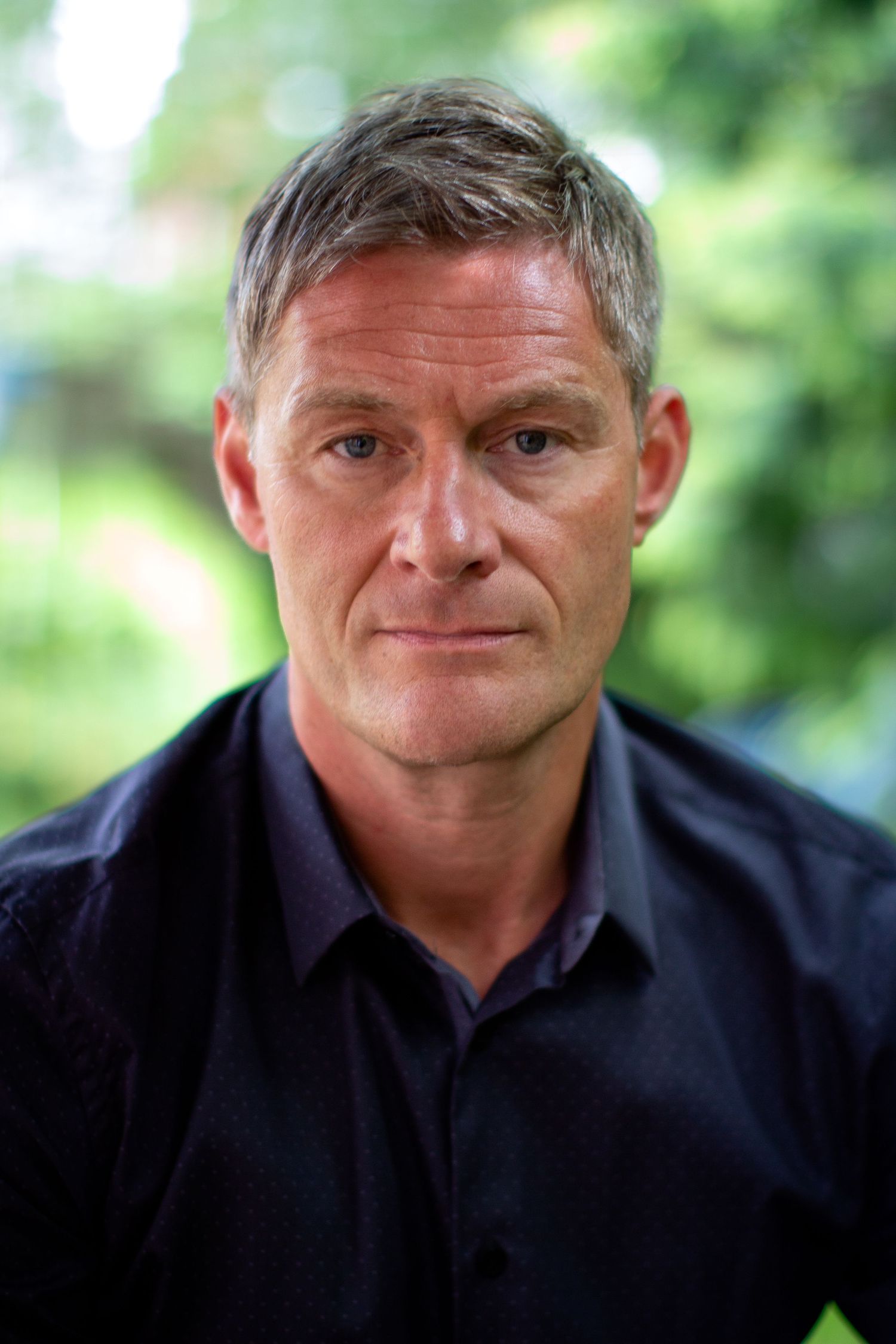 Colour portrait image of a client from for a headshot session. Client wearing a black shirt, contrasting the green textured outdoor setting.