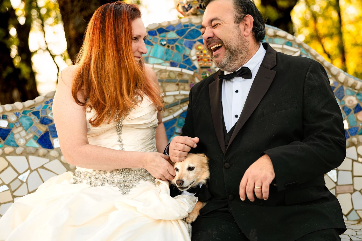 Bride and groom laughing together in wedding attire while holding a small dog on a mosaic bench outdoors.