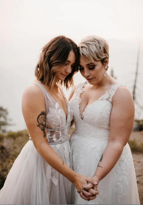 Two brides in white wedding gowns hold hands and touch foreheads in an intimate outdoor ceremony moment.