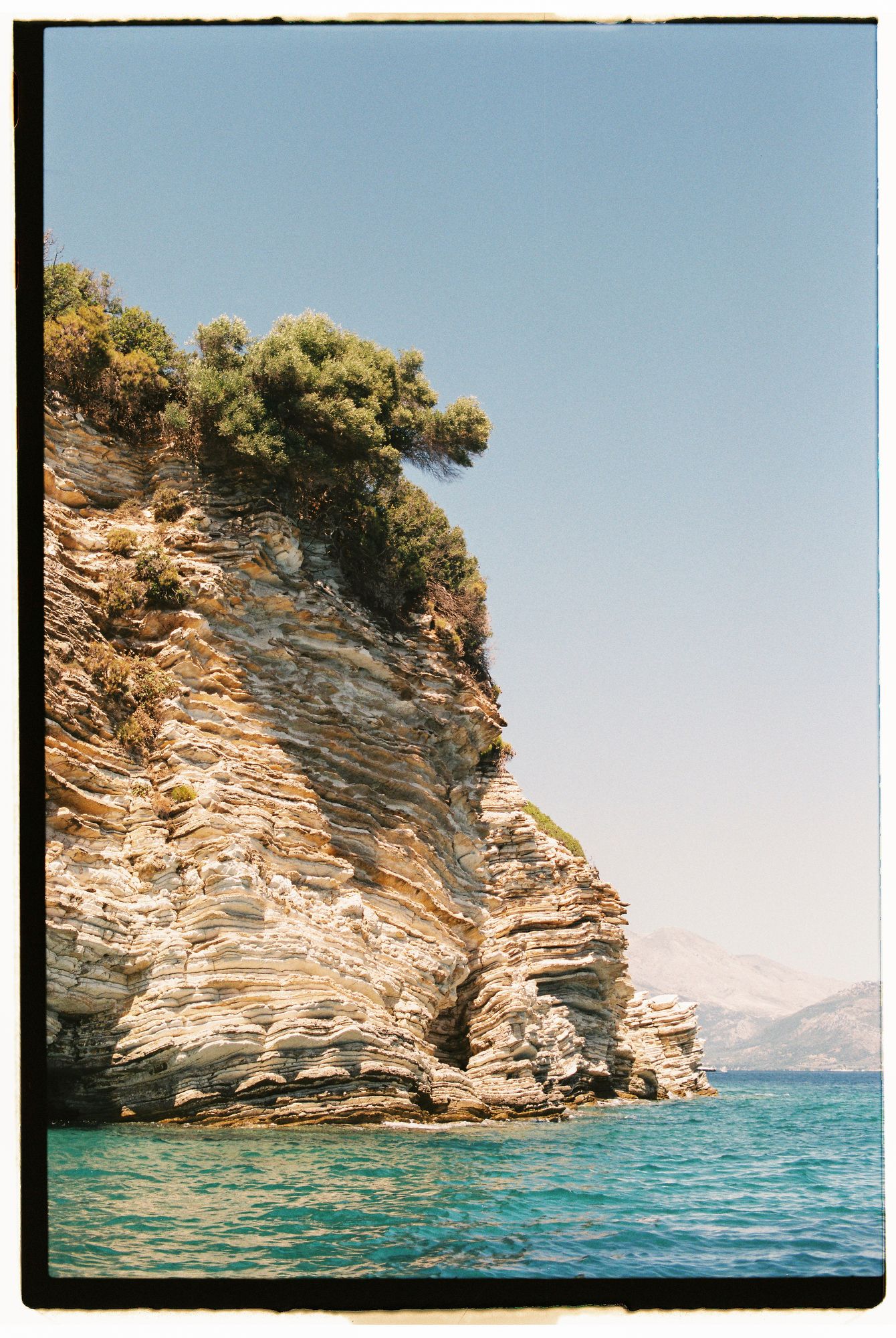 Rocky coastal cliffs rise dramatically from turquoise Mediterranean waters against a clear blue sky.