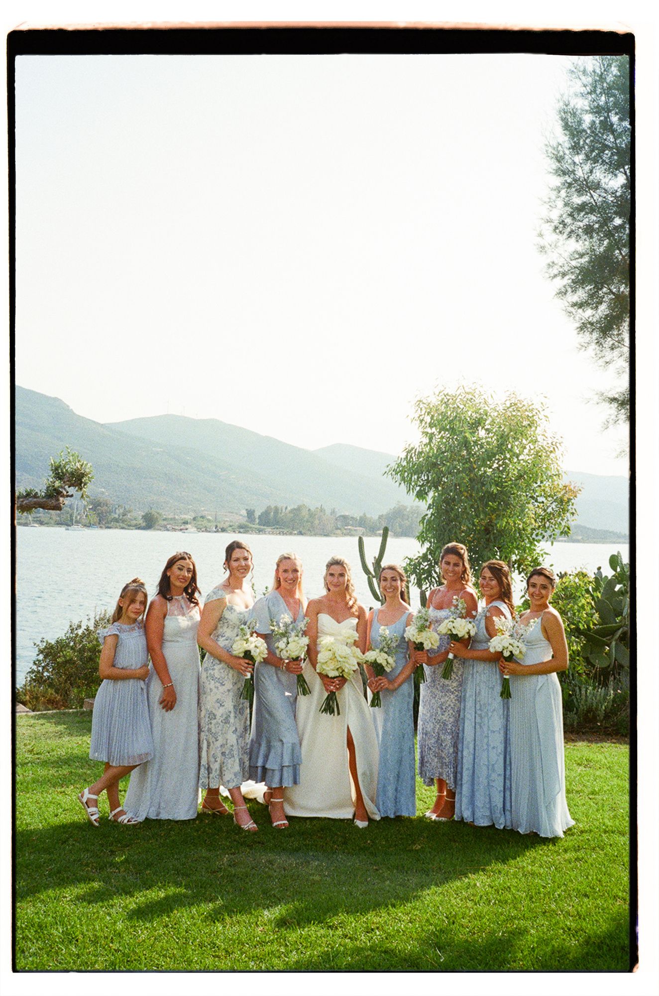 Bridesmaids in light blue dresses pose on a grassy hillside with mountain and lake views in the background.