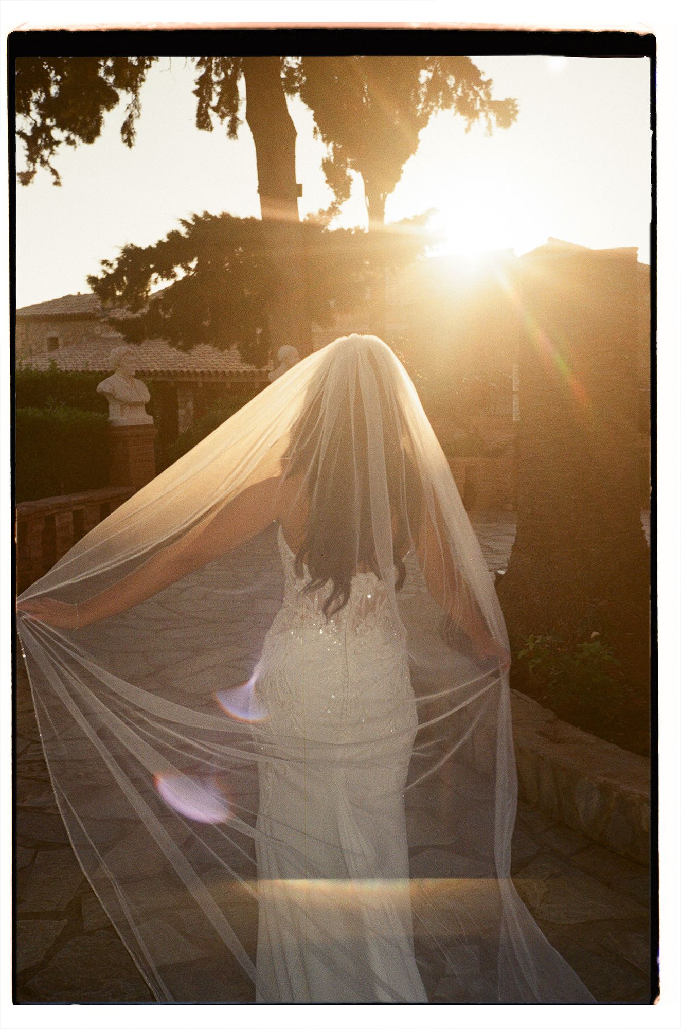 A bride in a flowing white wedding dress and veil stands silhouetted against a golden sunset.