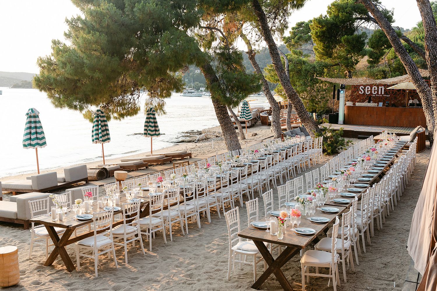Long rows of white chairs and tables set up for an elegant outdoor beach wedding reception under pine trees at Poros