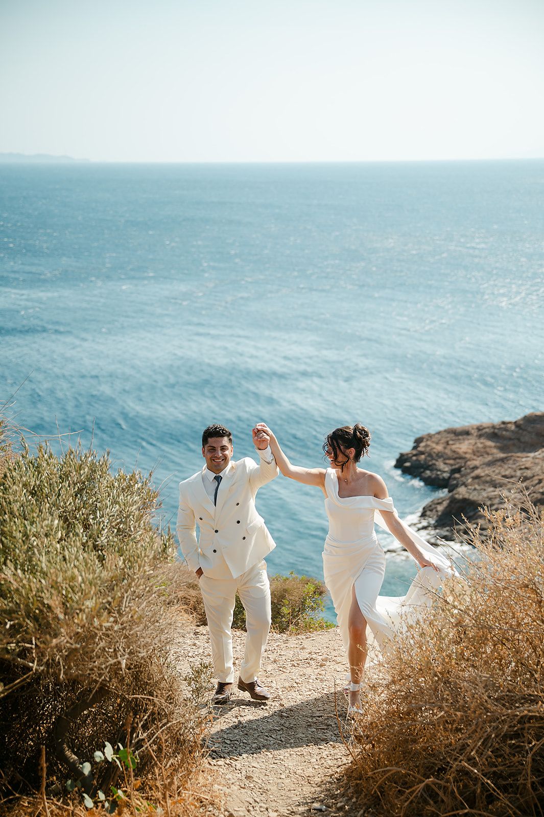 Couple in white outfits walking hand in hand along a scenic coastal cliff path with blue ocean views at Ktima 48 Athens
