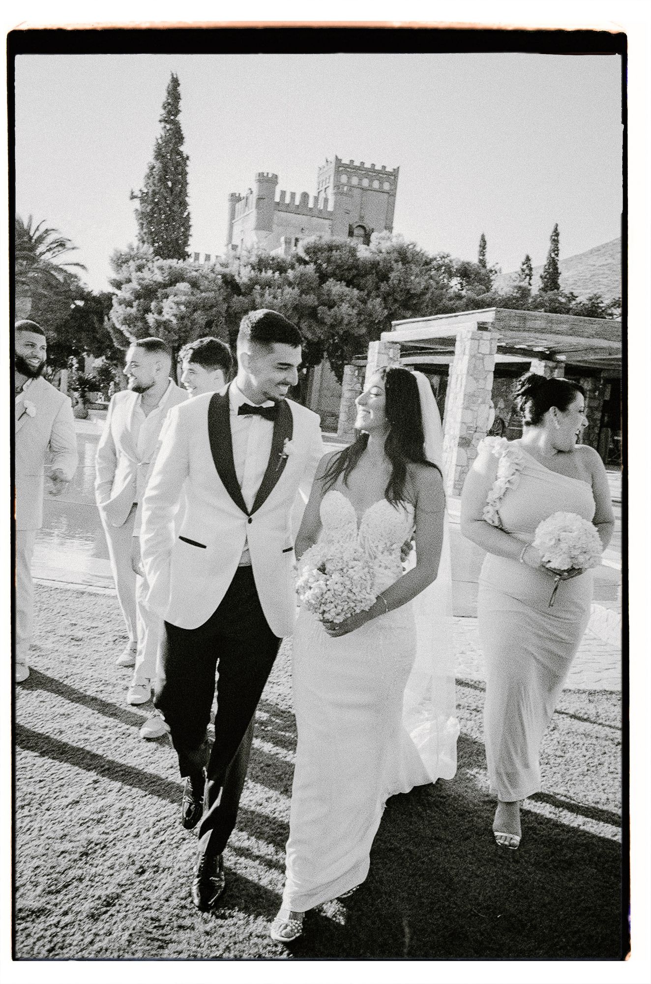 Wedding party walking together on gravel path near stone castle in black and white photography.