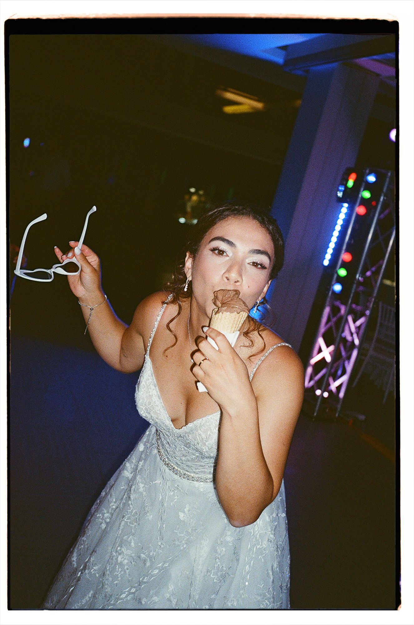A person in a white lace dress dances and poses playfully at a dimly lit party venue with neon lights in the background.