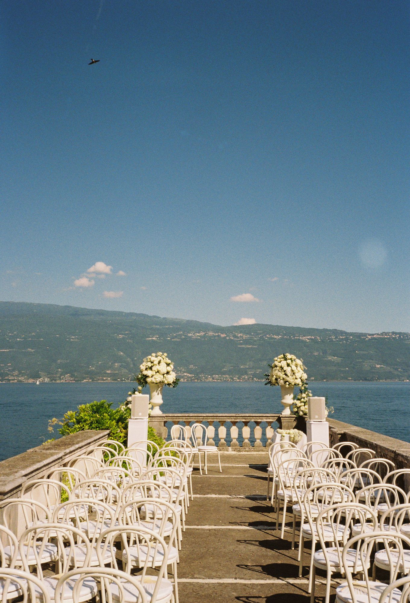 Elegant outdoor wedding ceremony setup at Lake Garda with white chairs and floral arrangements overlooking a scenic lake and mountains.