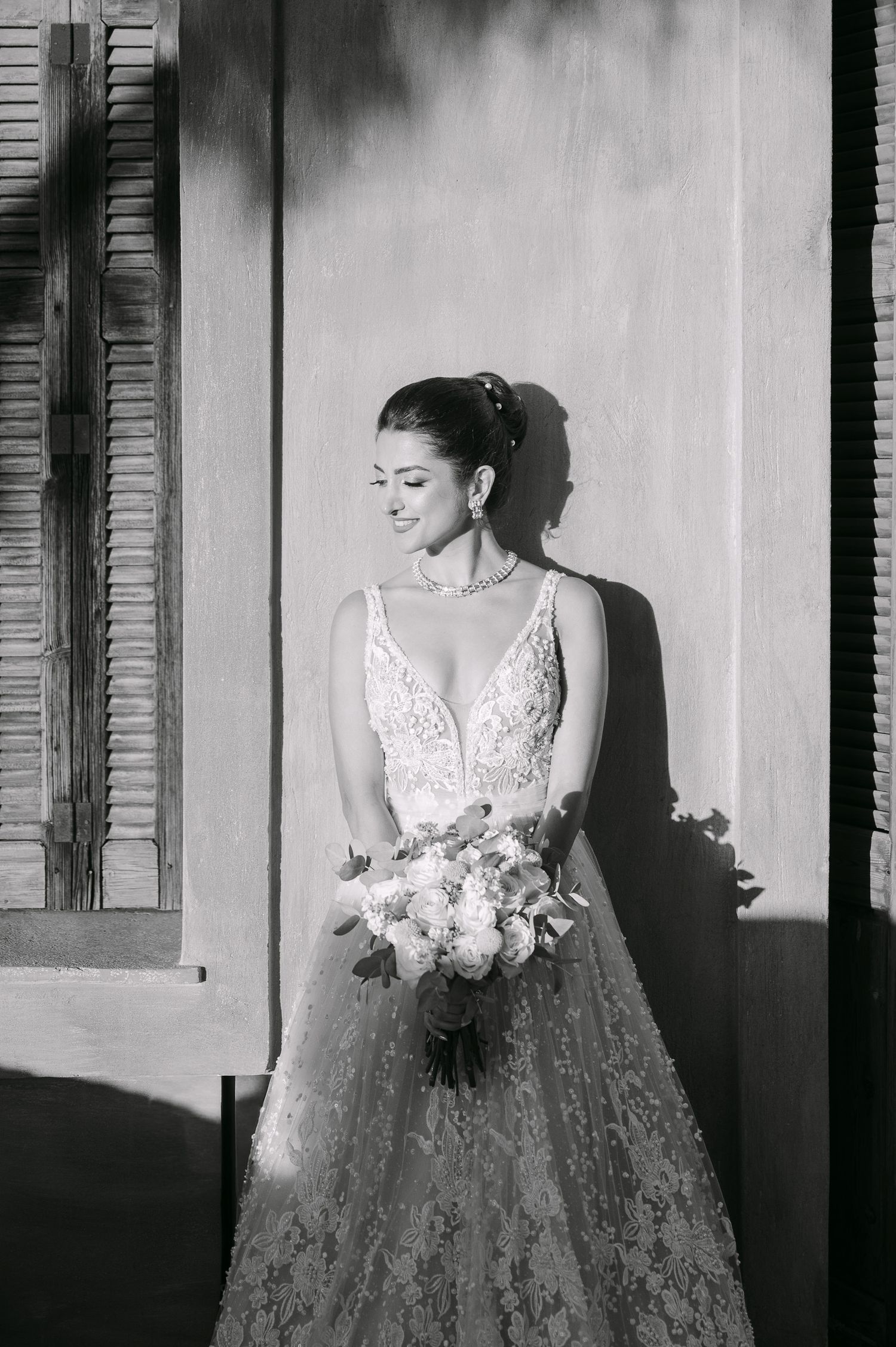 A black and white wedding portrait showing an elegant bride in a lace gown holding a floral bouquet near a window at Pyrgos Melissourgou