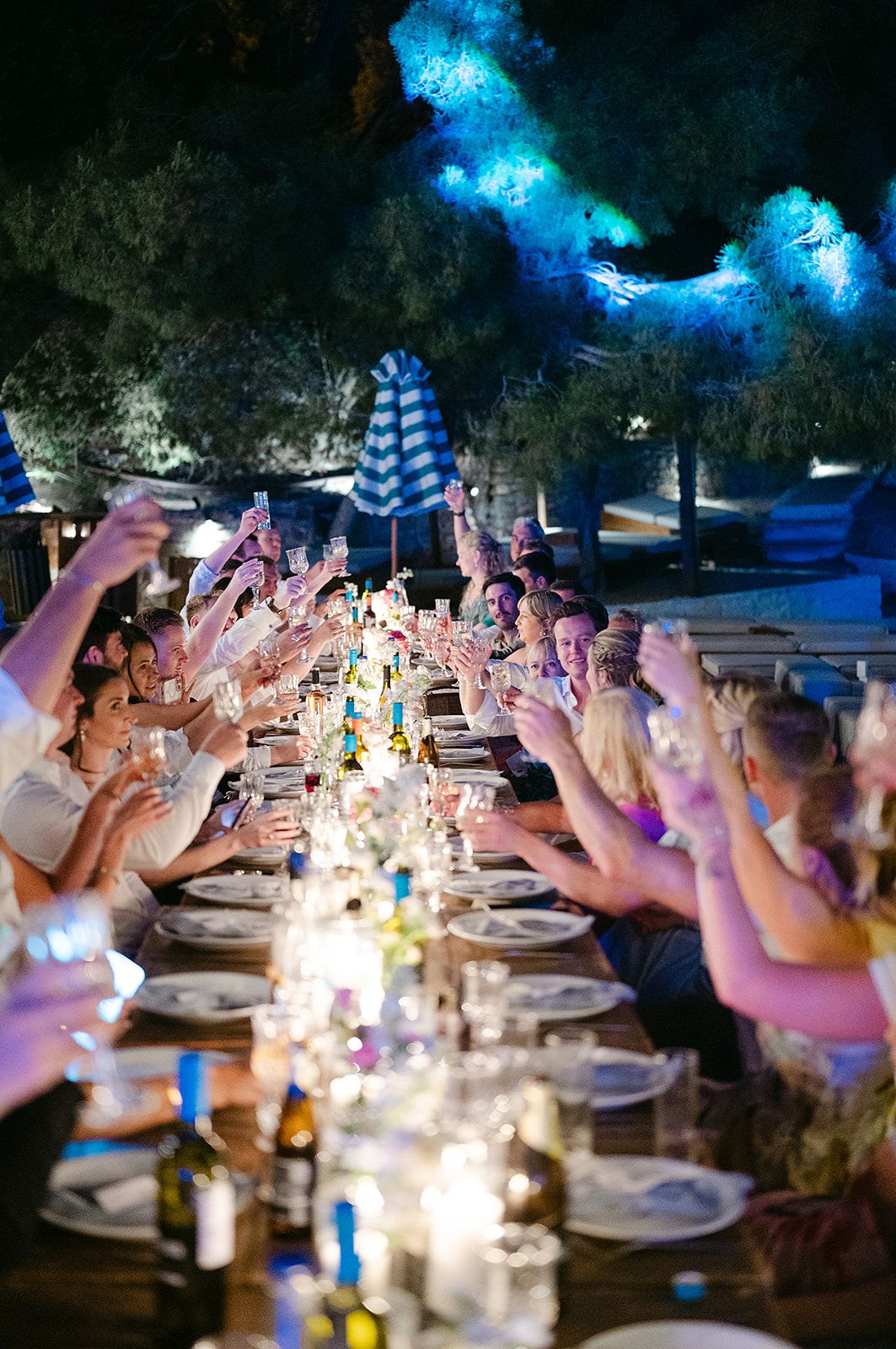 A candlelit outdoor dinner table with guests raising glasses in a toast under blue-lit trees during an evening celebration at Poros