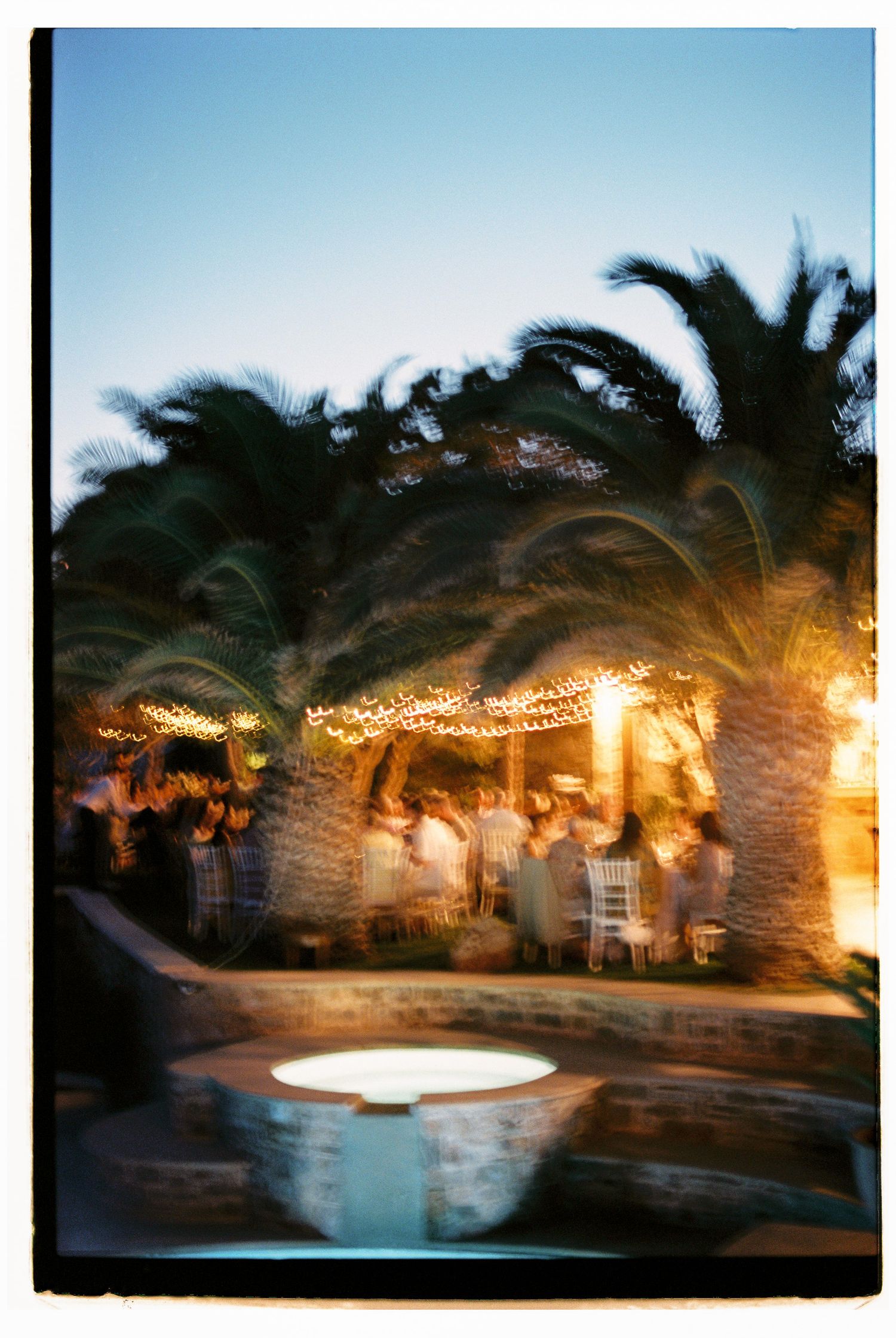 String lights illuminate a tropical poolside patio with palm trees and stone walls at dusk creating a romantic ambiance at Cycladic Gem Ios