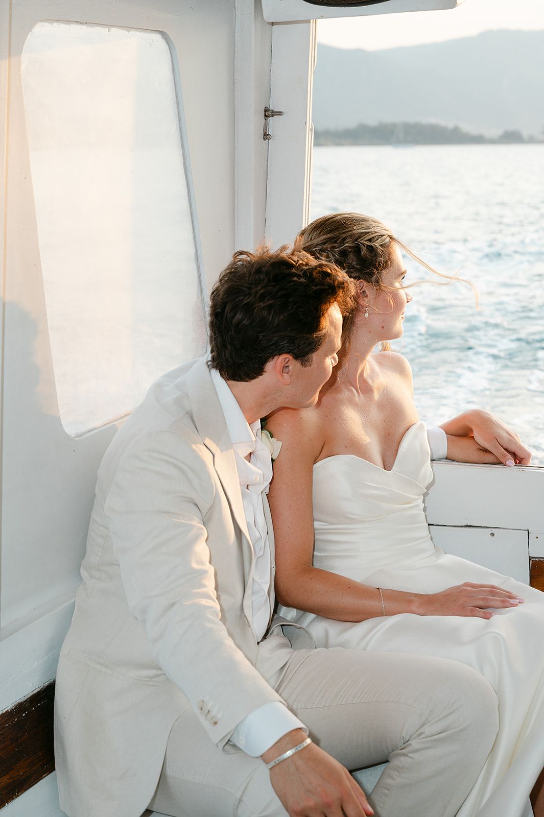 A couple in white wedding attire sits together on a boat deck overlooking the ocean on their special day at Poros