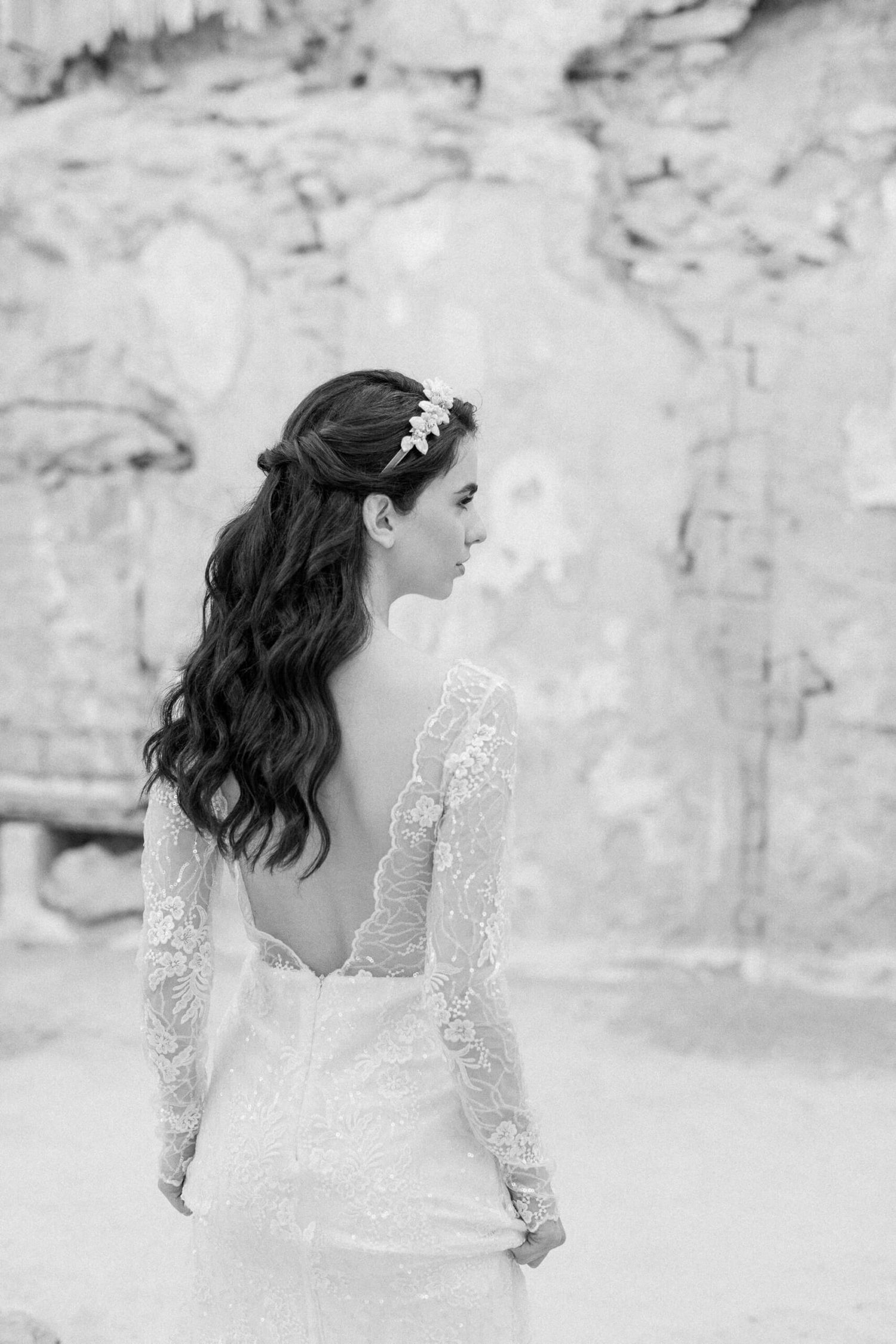 A bride in a white lace wedding dress with long sleeves poses against a textured stone wall in black and white.
