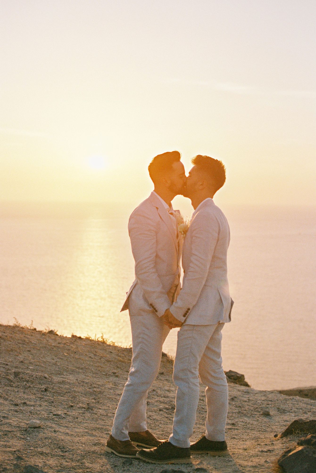 A romantic couple in white attire shares an intimate moment at sunset on a coastal cliff overlooking the sea at Santorini.