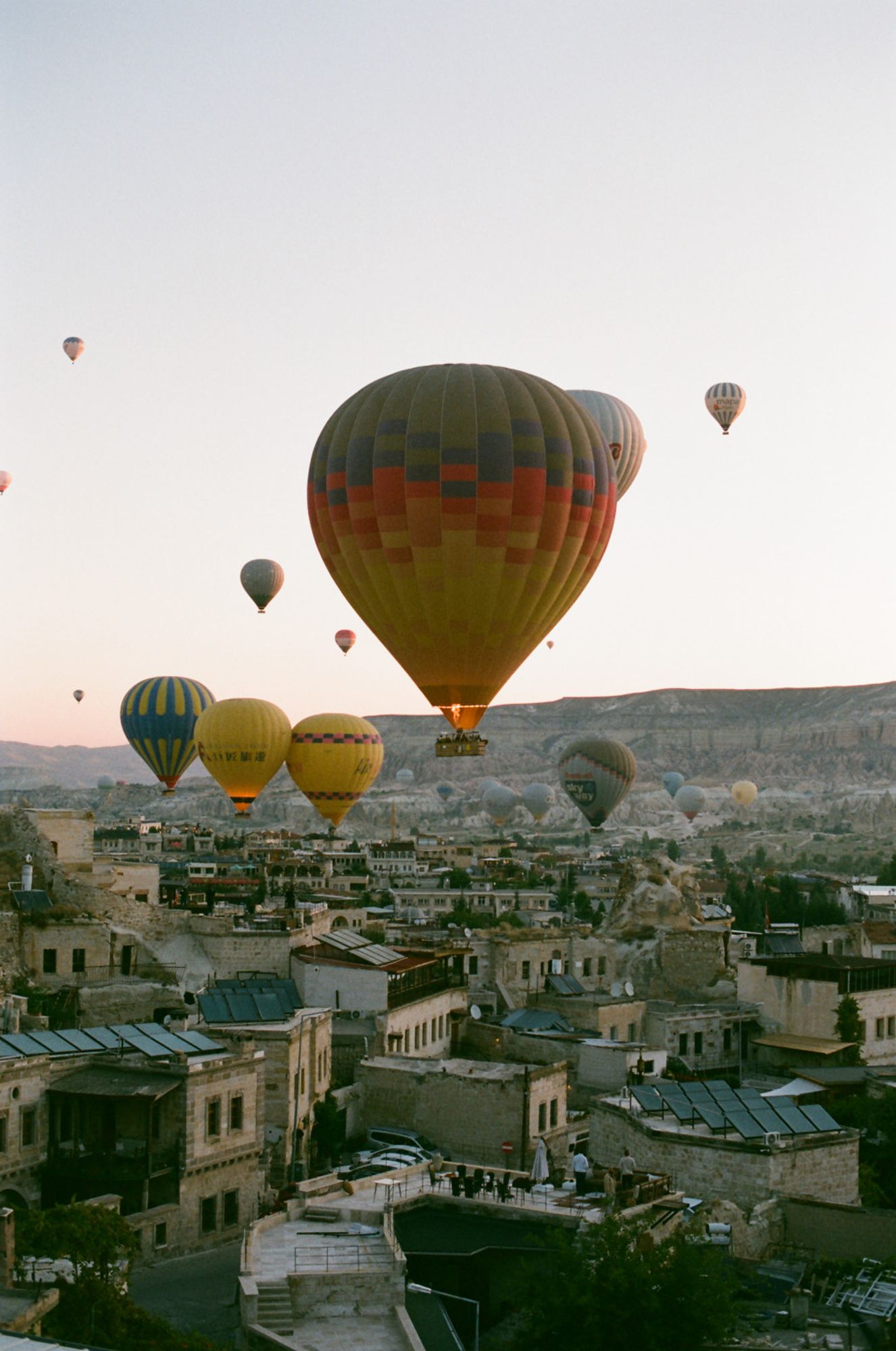 Hot air balloons float over ancient stone buildings in Cappadocia, Turkey during sunrise with warm morning light.