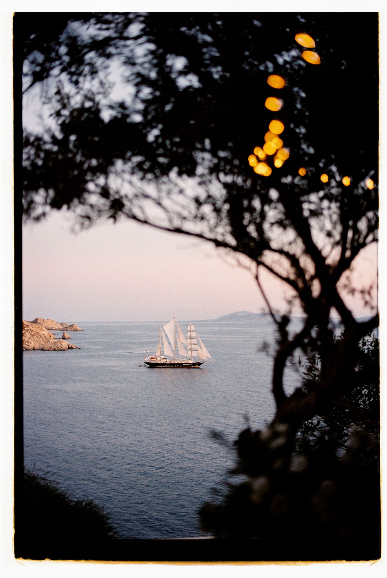 A sailing ship silhouetted against the Mediterranean Sea at dusk, framed by dark tree branches and glowing lanterns.