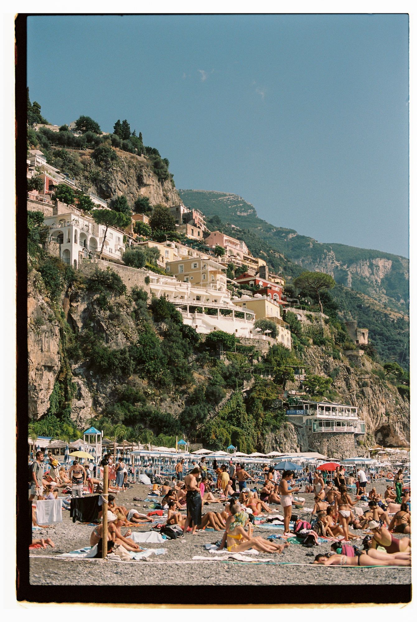 Crowded beach along the Amalfi Coast with colorful umbrellas and cliffside buildings perched on steep mountainside.