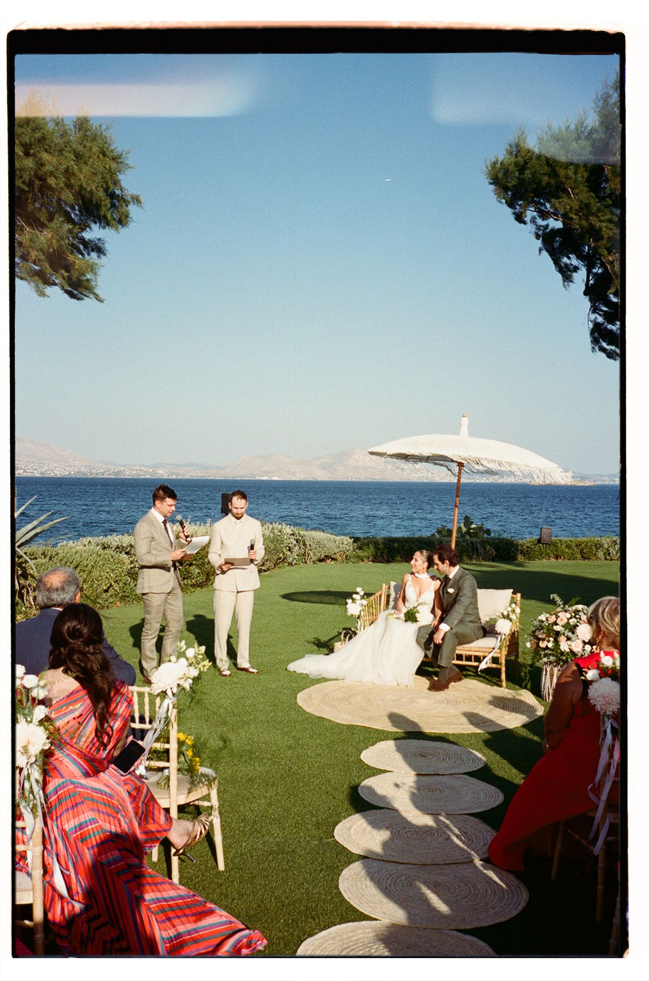 Outdoor wedding ceremony setup on a grassy oceanfront lawn with white chairs and parasols under blue skies.