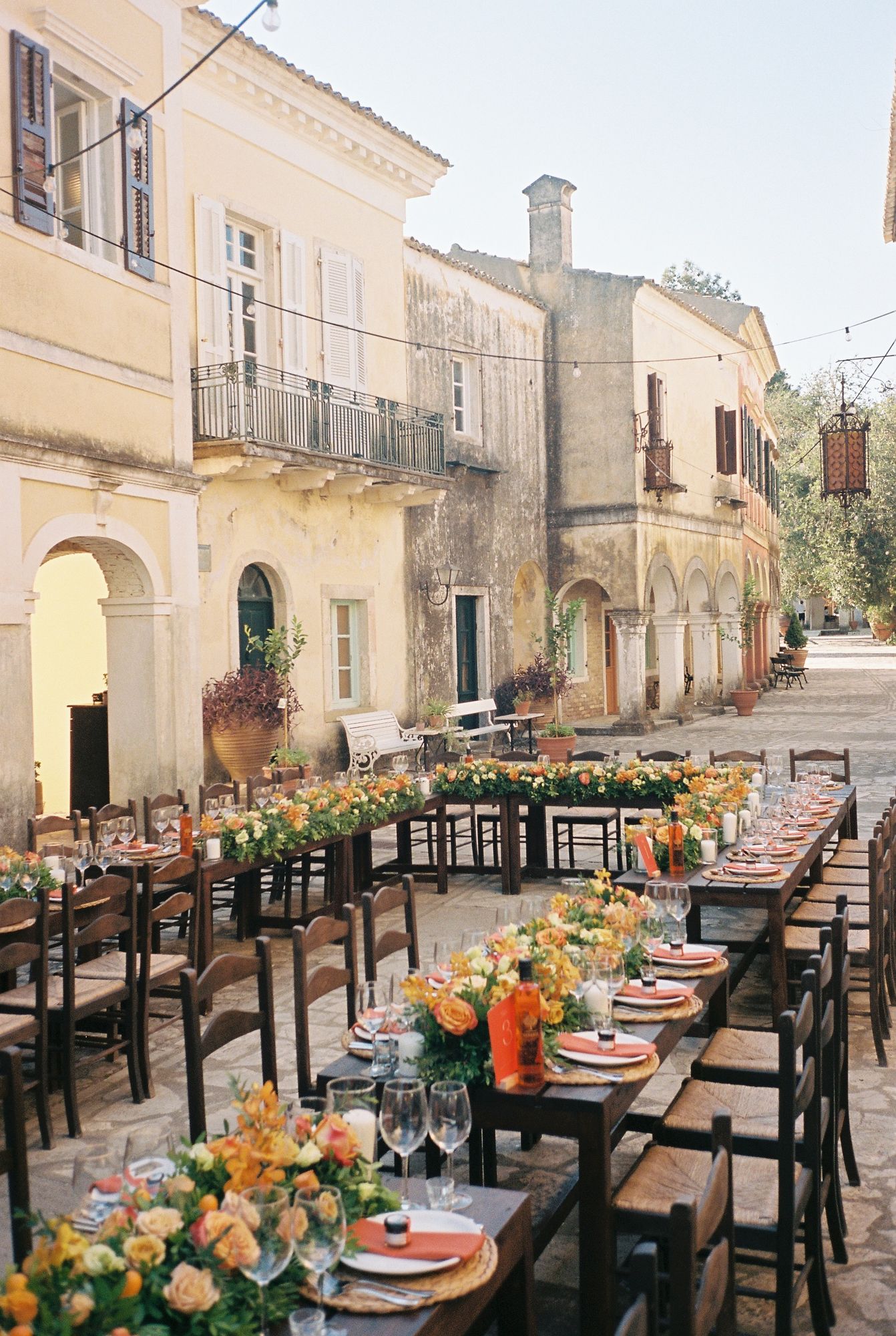 Long rustic wooden tables set for an elegant outdoor dinner party with orange floral centerpieces and place settings.