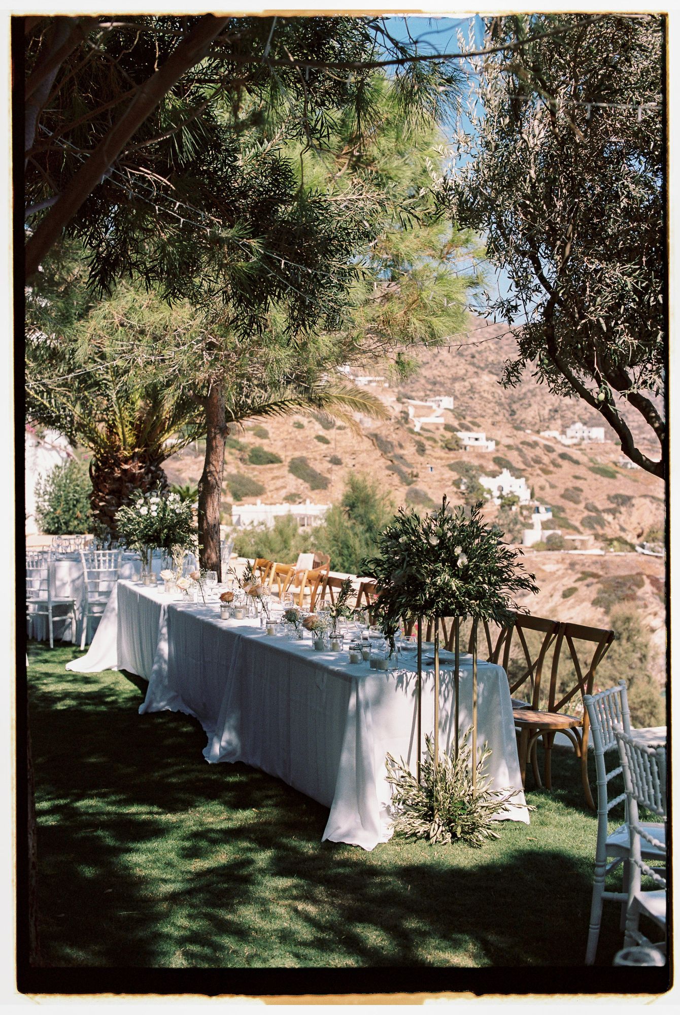 Elegant outdoor wedding reception table with white linens set against a scenic hillside backdrop under shaded trees.