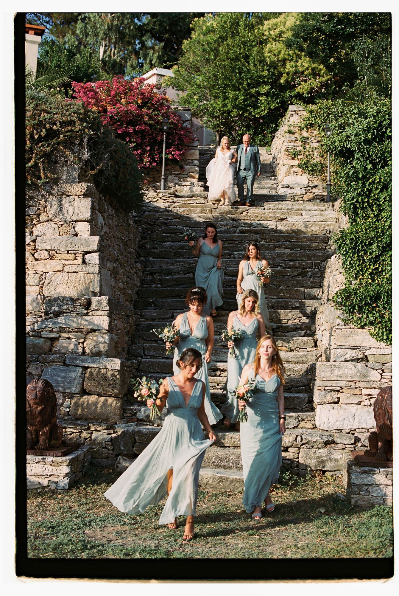 Wedding party in light blue dresses descends stone steps in a garden setting during an outdoor celebration.