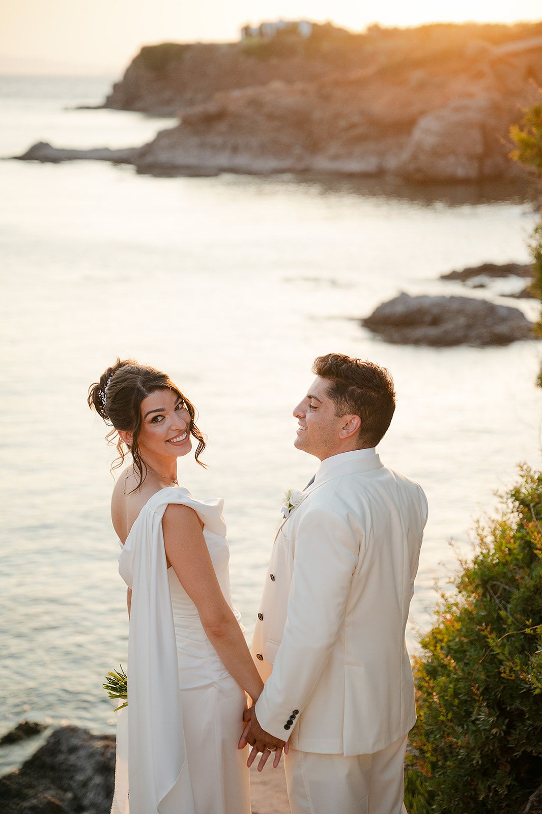 A couple in white attire shares a romantic moment at sunset by the ocean cliffs at Ktima 48 Athens