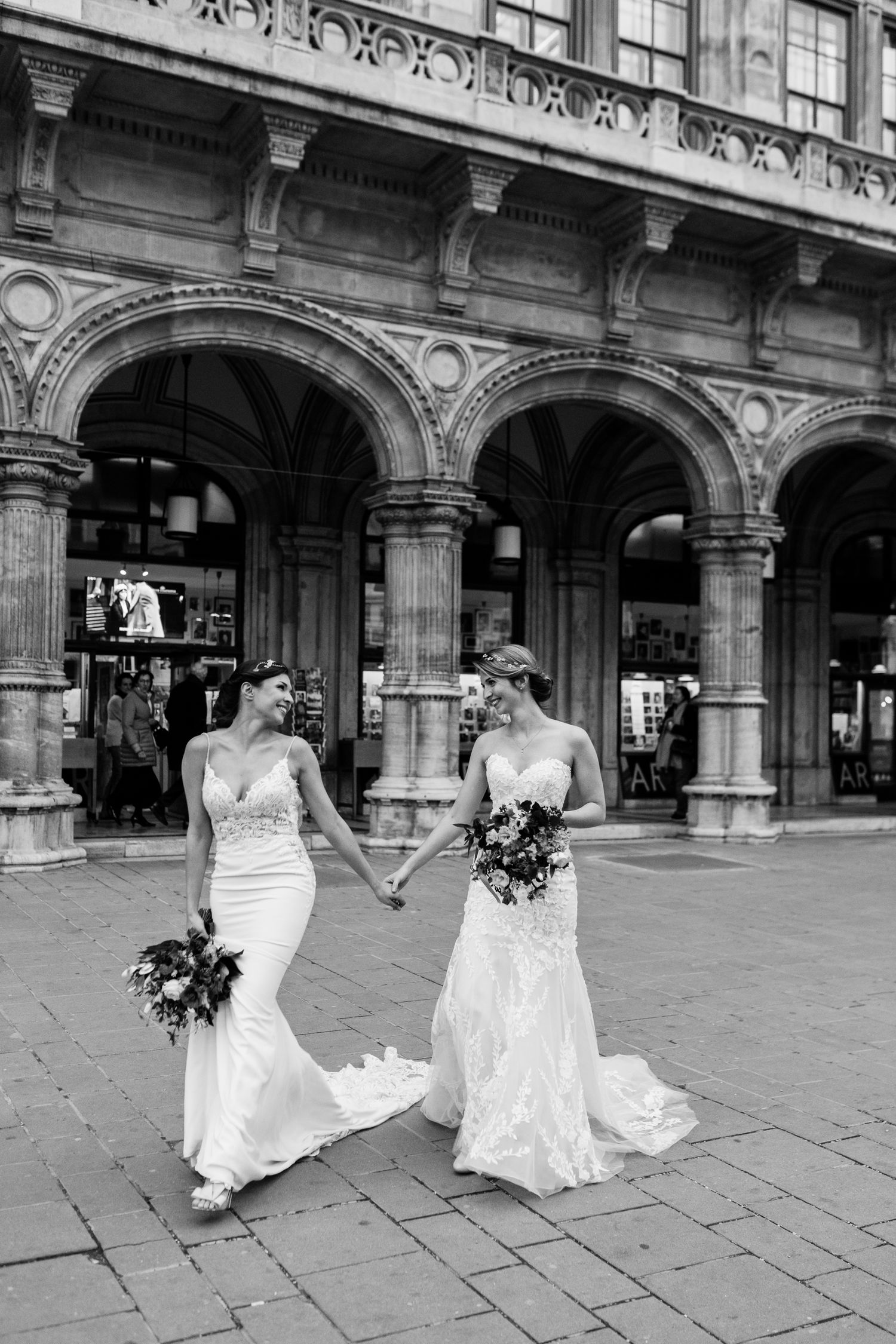 Two brides in elegant white lace wedding dresses hold hands while walking under ornate stone archways in black and white at Vienna Austria