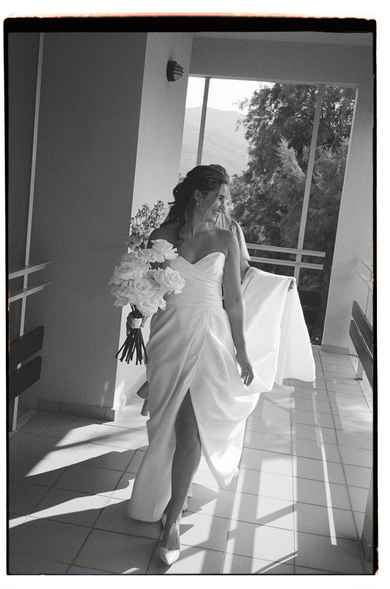 A bride in a flowing white gown stands in sunlit doorway holding a bouquet on her wedding day.