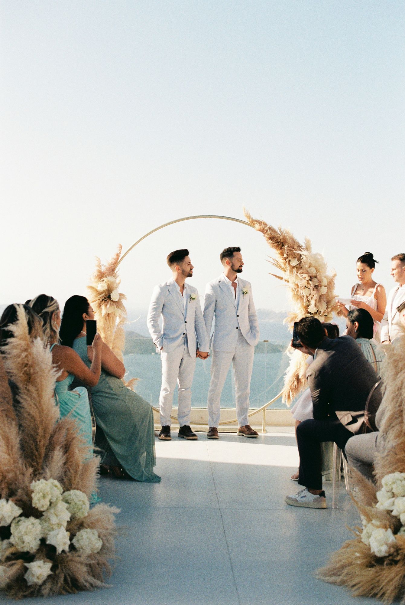 Wedding guests in white and dark attire gather on an outdoor terrace with pampas grass and floral decorations at El Viento Santorini